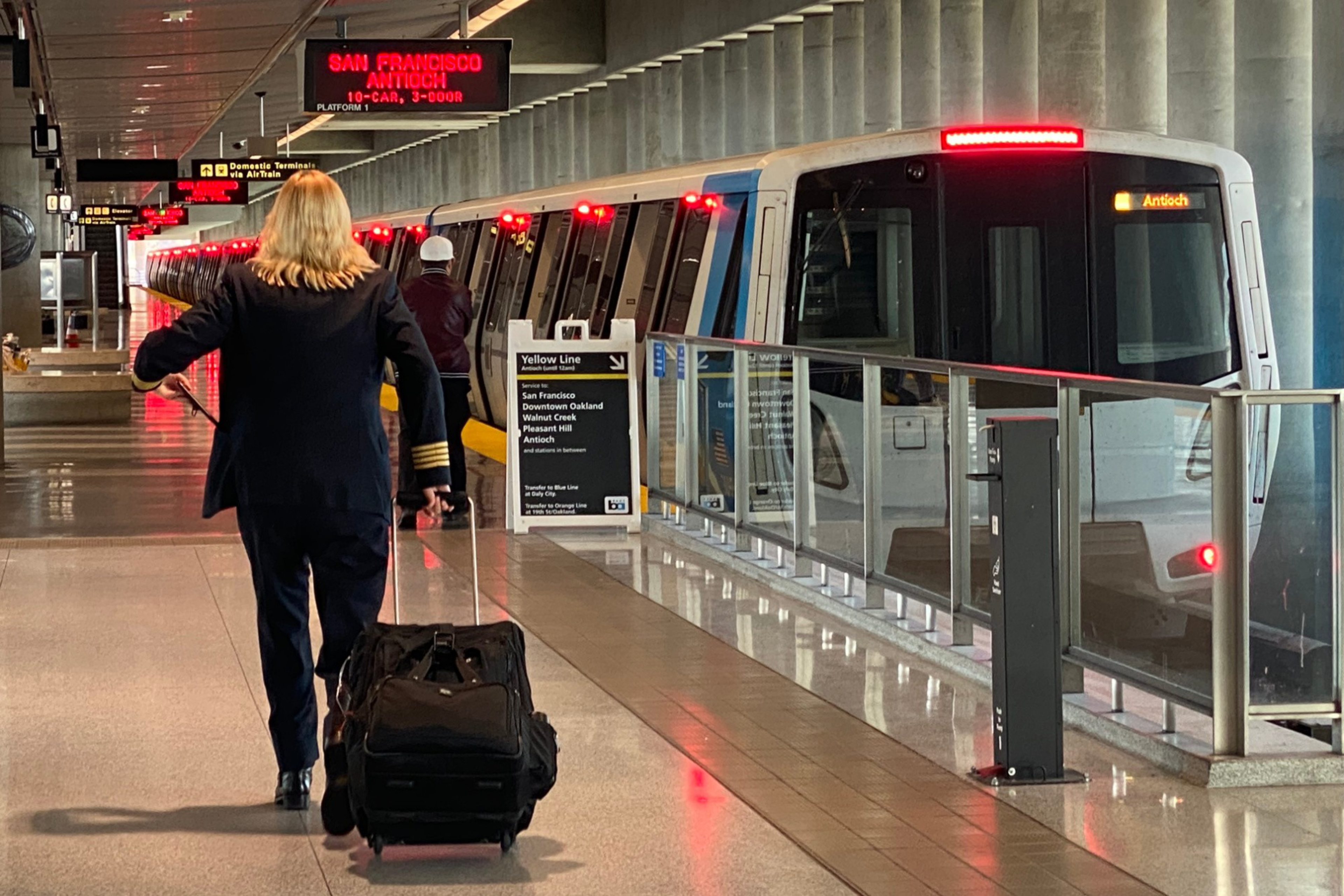 A person in a uniform pulls a suitcase along a terminal platform next to a stationary train with red lights and signs for San Francisco and Antioch.