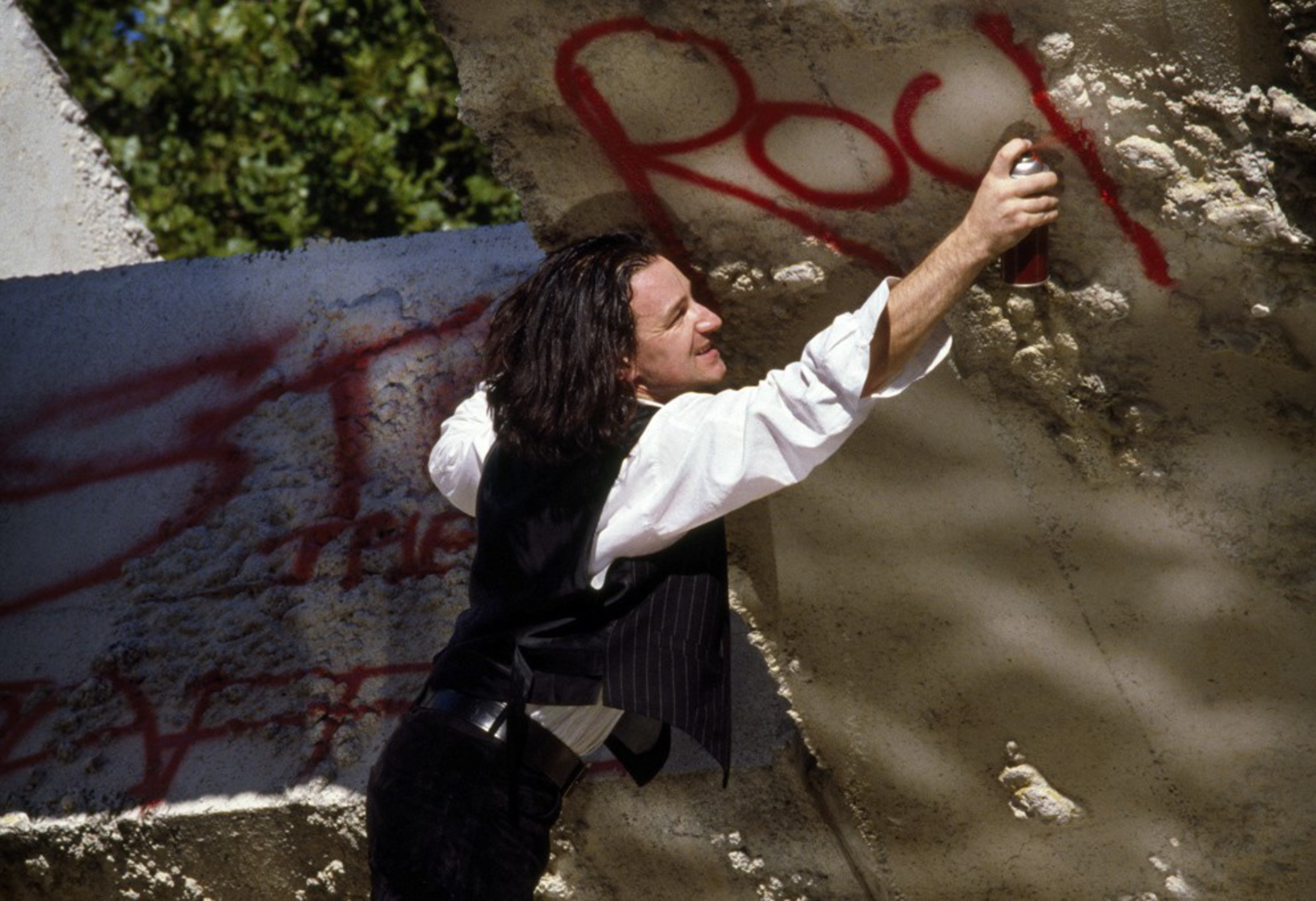 A person with long hair spray-paints the word &quot;Rock&quot; in red on a textured concrete wall. They wear a white shirt and black vest. Trees are visible in the background.