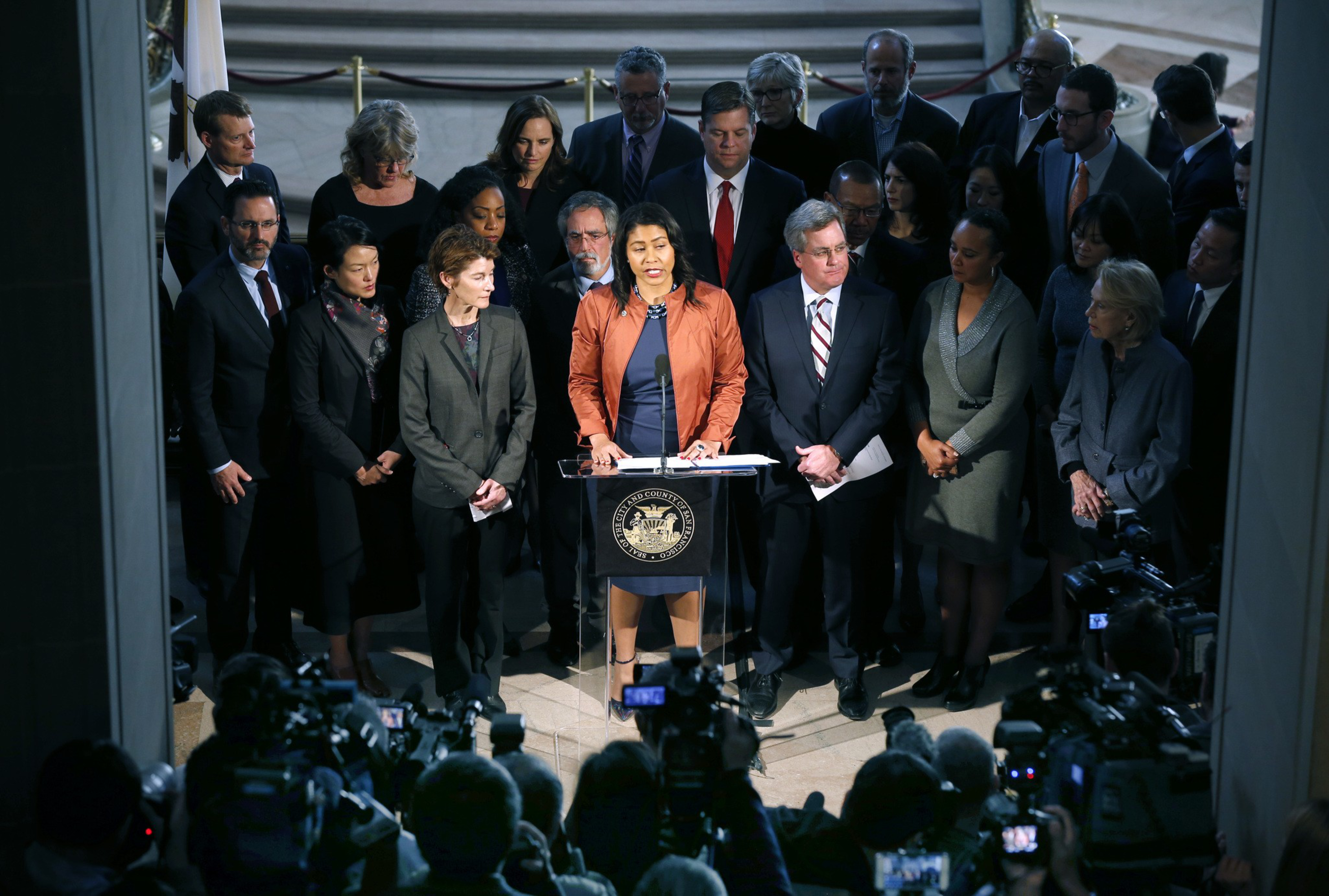 A woman speaks at a podium surrounded by a large group of people. Cameras capture the event in a formal setting with a government emblem on the podium.