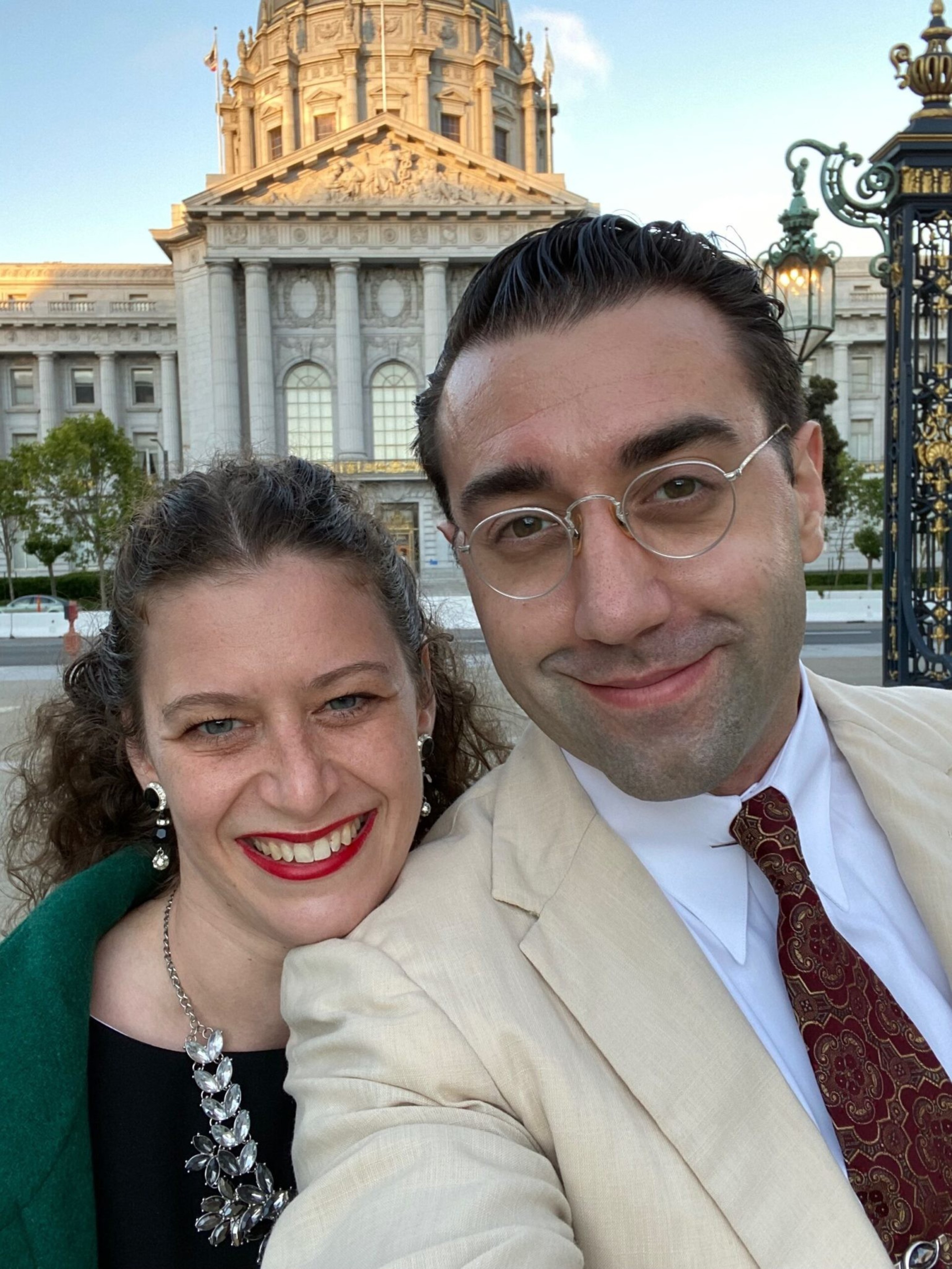 A smiling man in glasses and beige suit stands next to a woman with red lipstick and curly hair, with a grand classical building behind them.