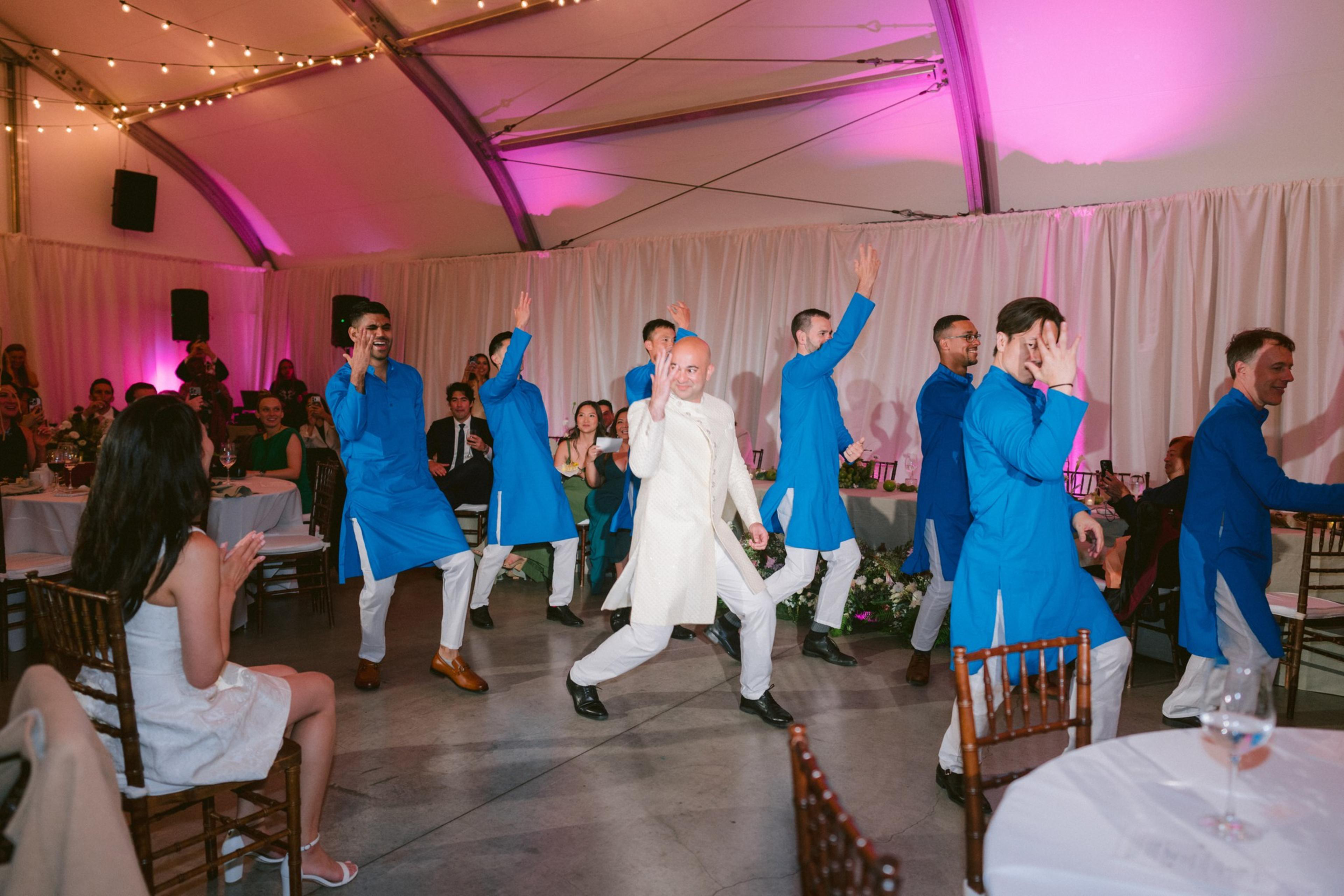 A group of men in blue and white traditional outfits perform a coordinated dance in front of seated guests applauding, under pink and white decorated lighting.