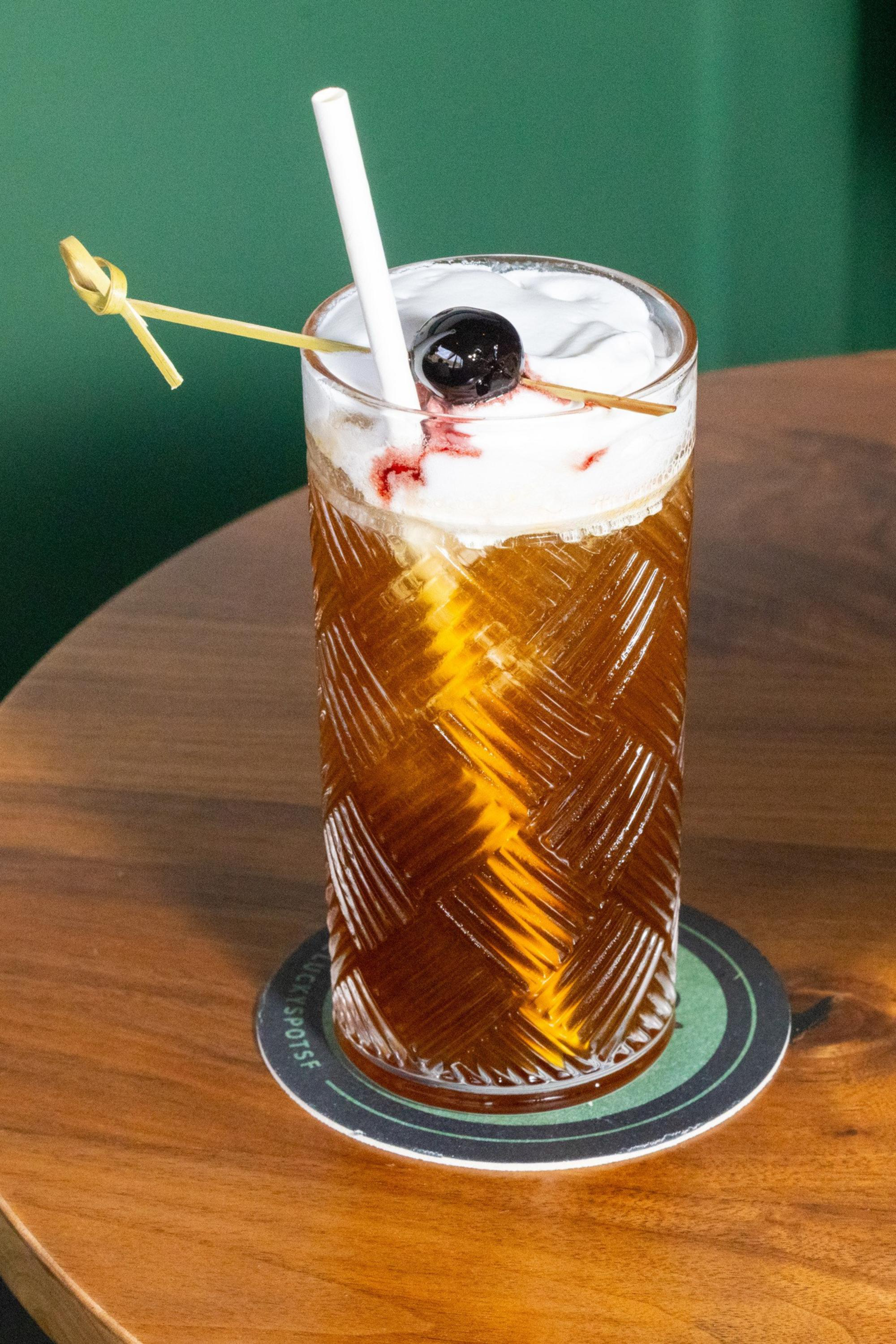 A tall, textured glass filled with an iced brown drink, topped with foam and a dark cherry on a skewer, with a white straw inside, sits on a wooden table.
