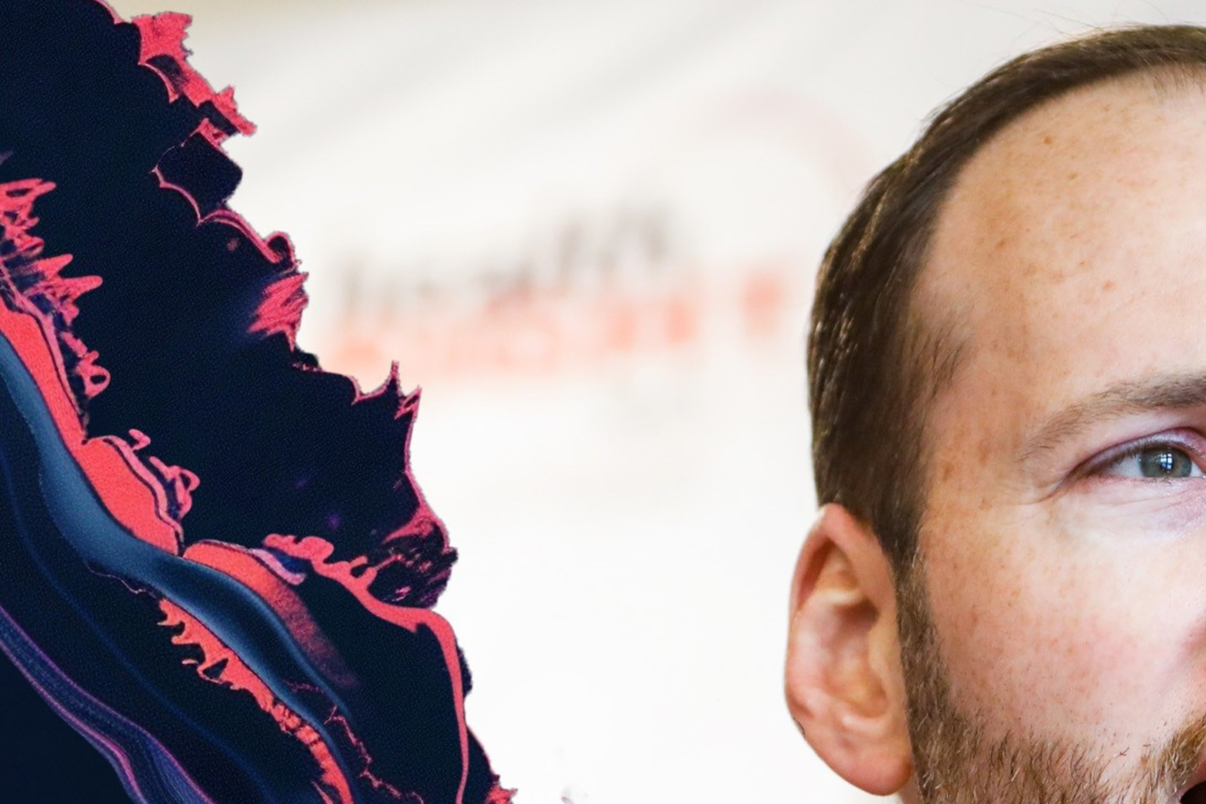A man in a suit and tie speaks, looking to the side. He has a beard, and there's a colorful, abstract wave pattern surrounding the image.