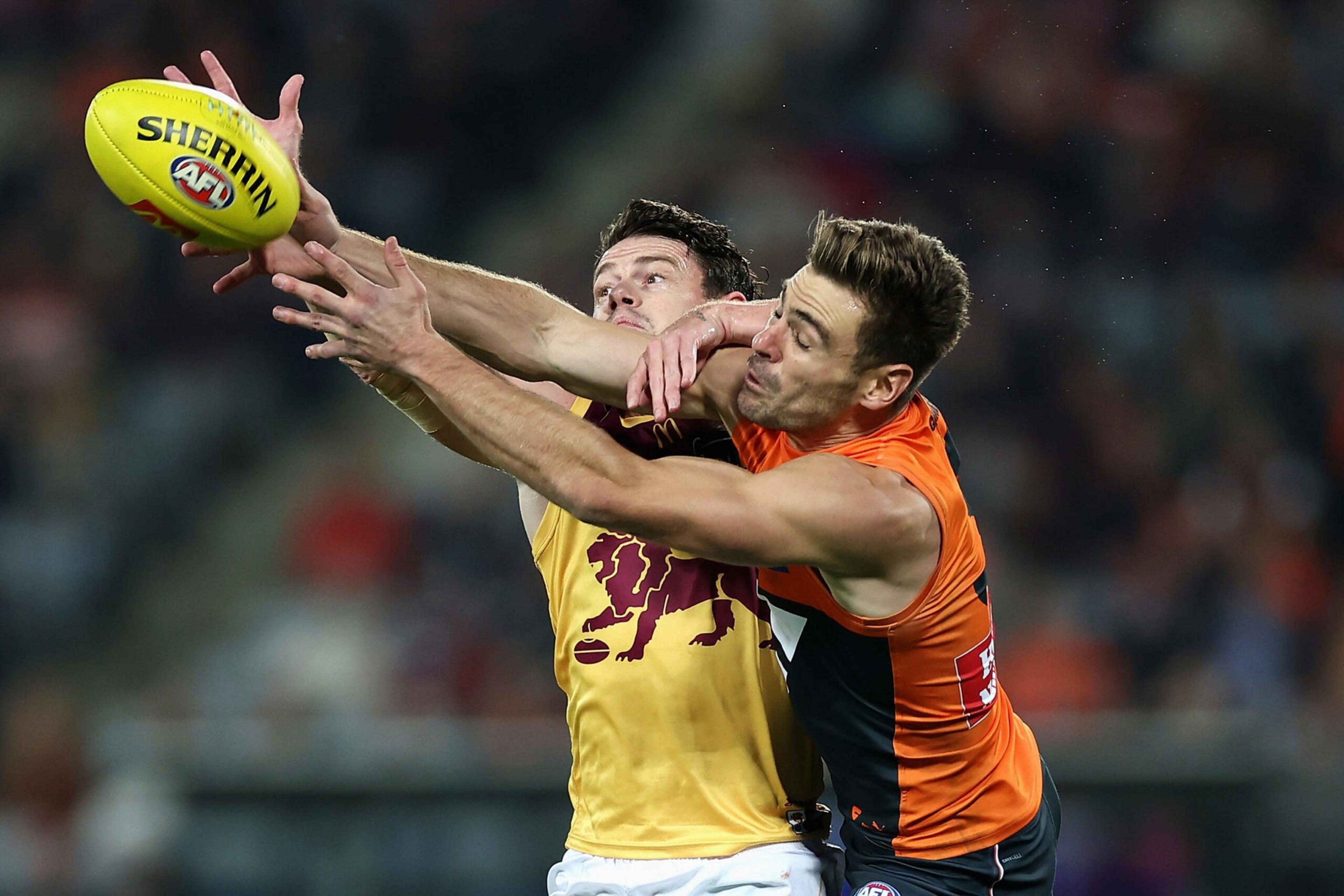 Two Australian football players in yellow and orange jerseys fiercely compete to catch a yellow Sherrin football during a game.