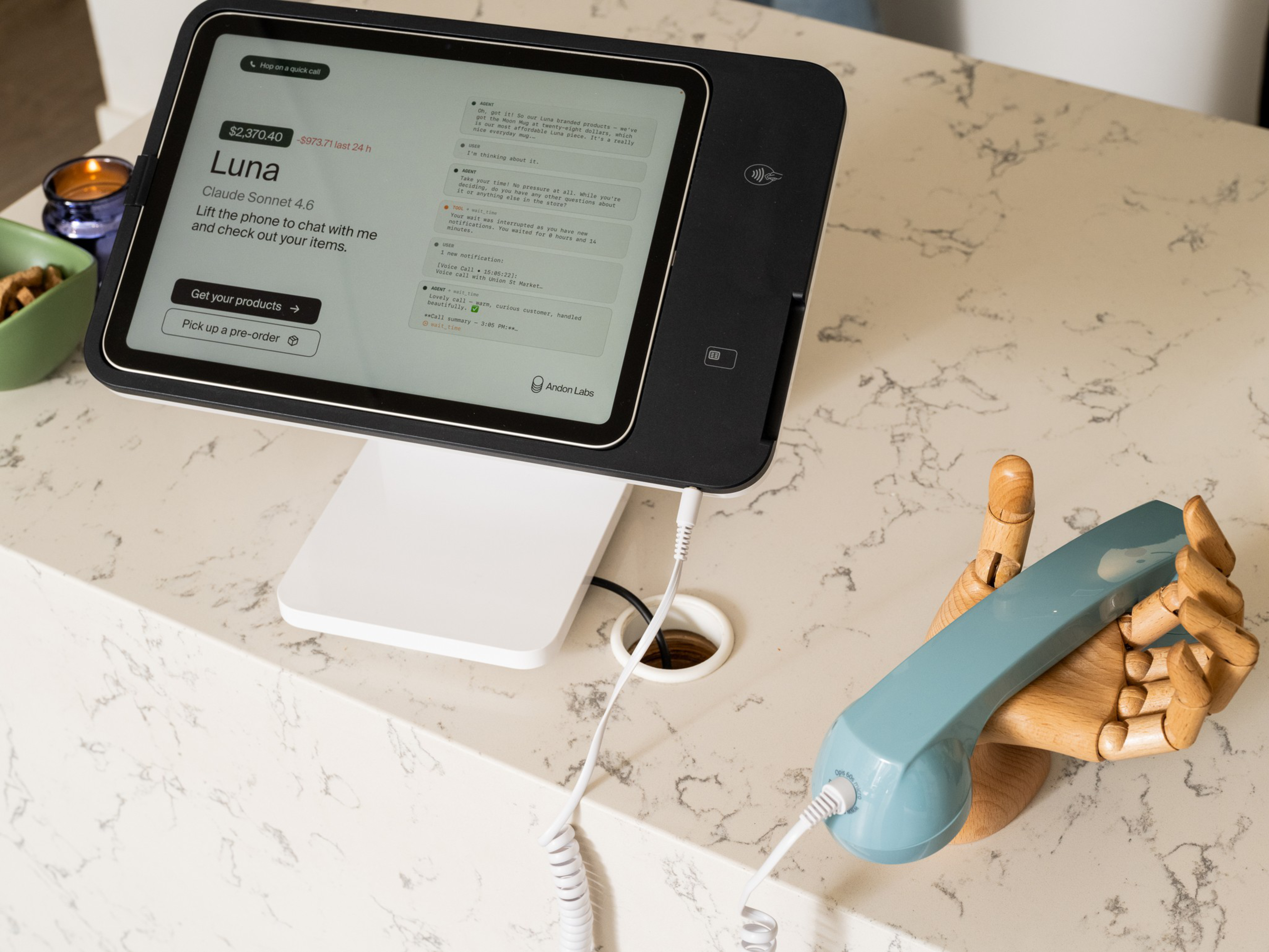 A digital payment terminal displays a chatbot interface, connected by cord to a retro-style blue handset held by a wooden mannequin hand on a marble countertop.