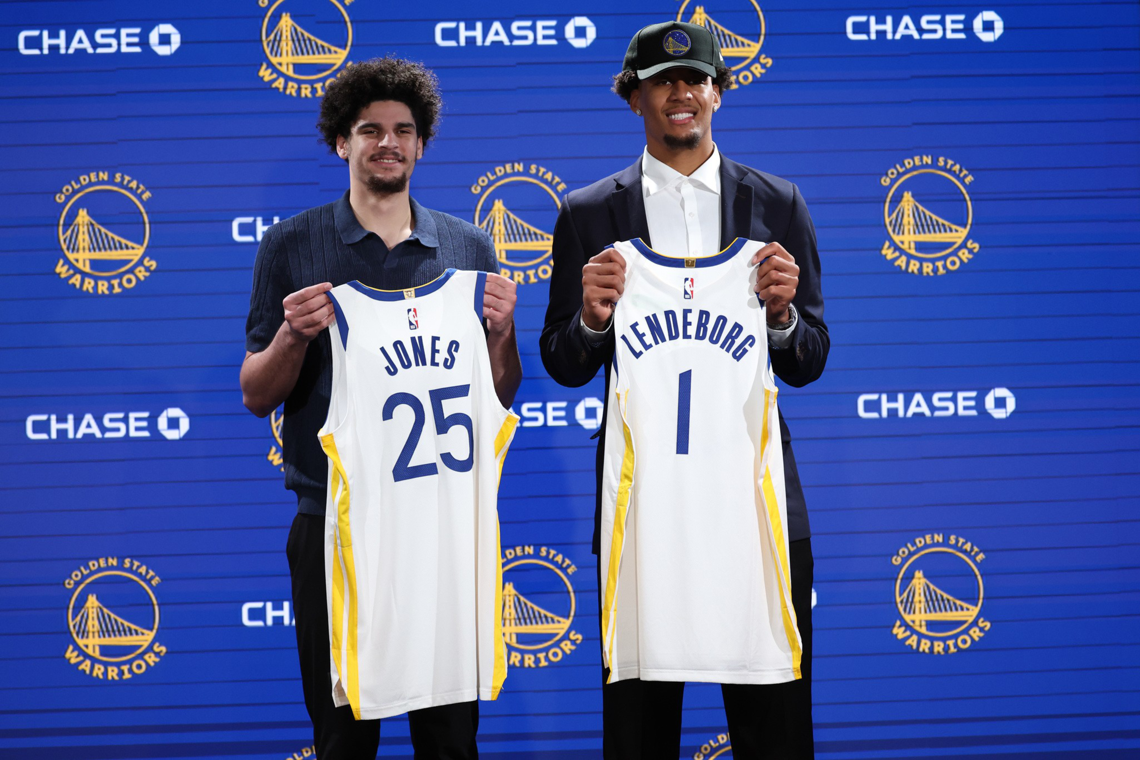 Two men stand in front of a Golden State Warriors backdrop, each holding a Warriors jersey with their last names and numbers visible.