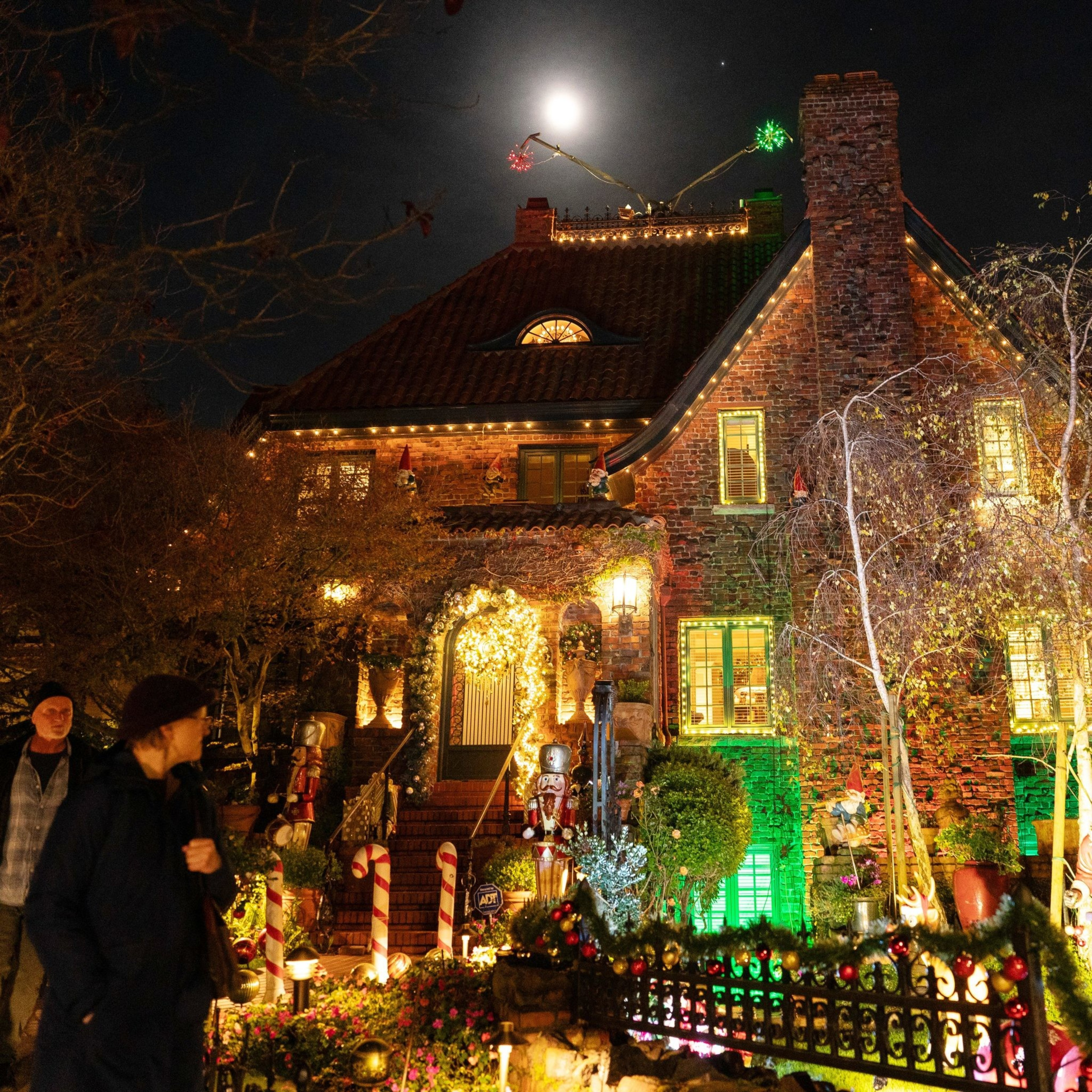 A brick house is festively decorated with Christmas lights and ornaments. The garden has candy cane decorations, and the moon shines brightly overhead.