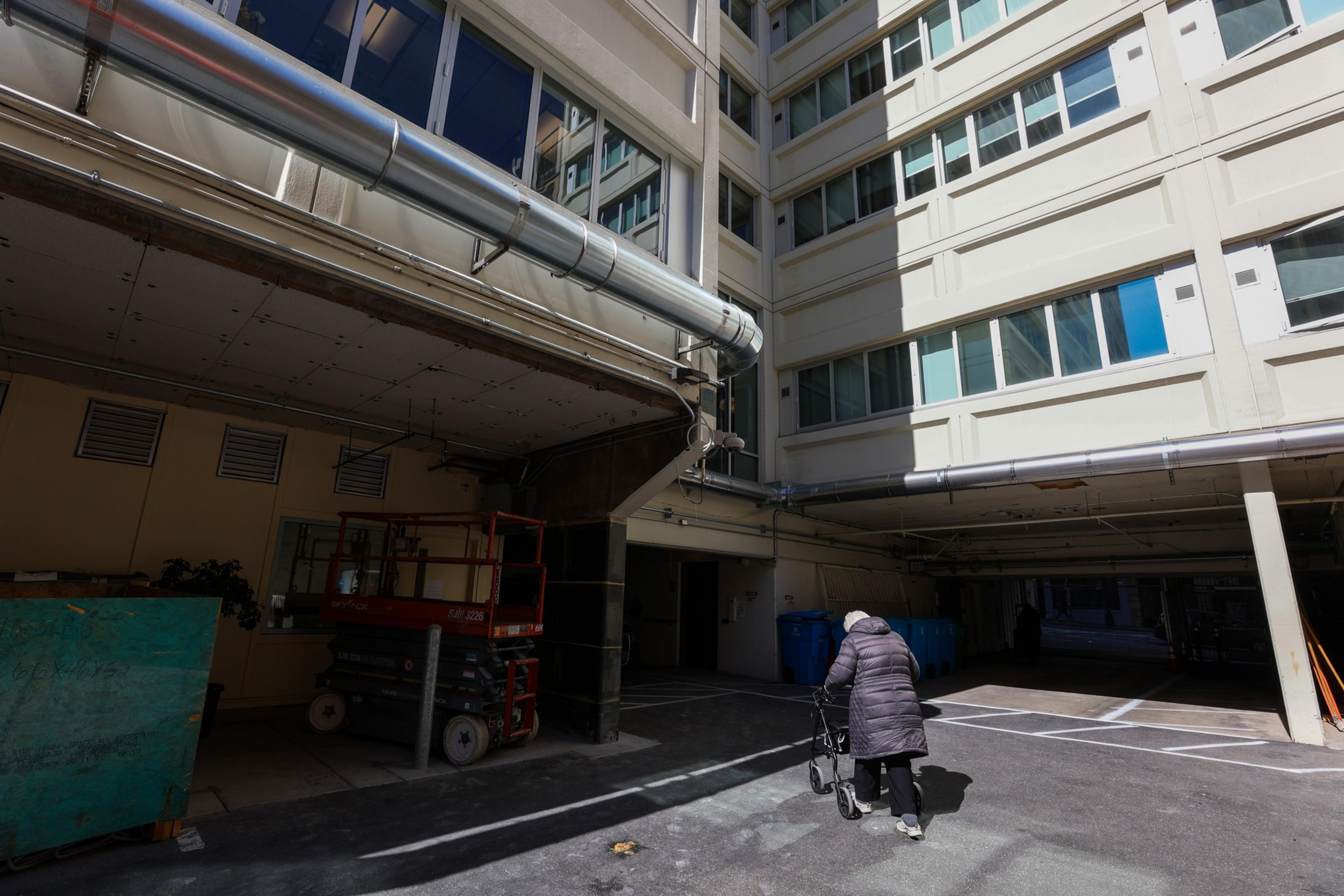 An elderly person using a walker moves through a shadowed courtyard between tall buildings with multiple windows and visible pipes overhead.
