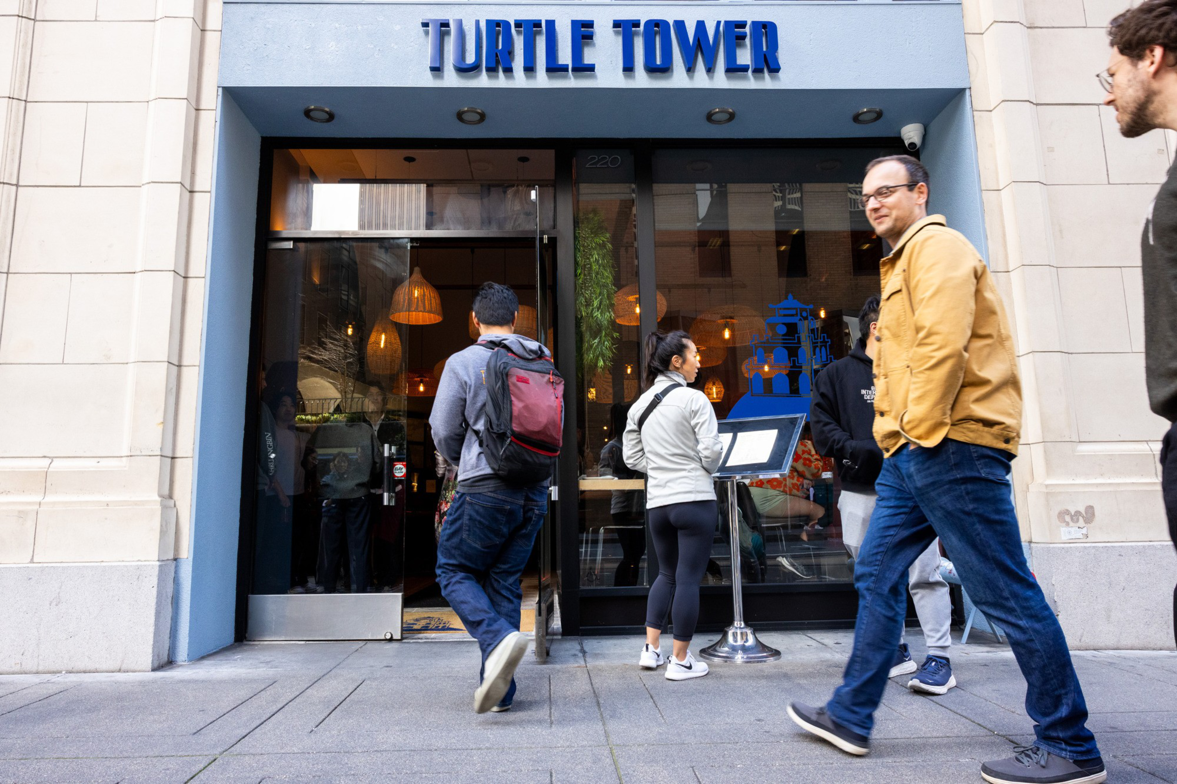 A restaurant entrance labeled "Turtle Tower" with a blue frame has people walking by and a woman reading the menu displayed outside.