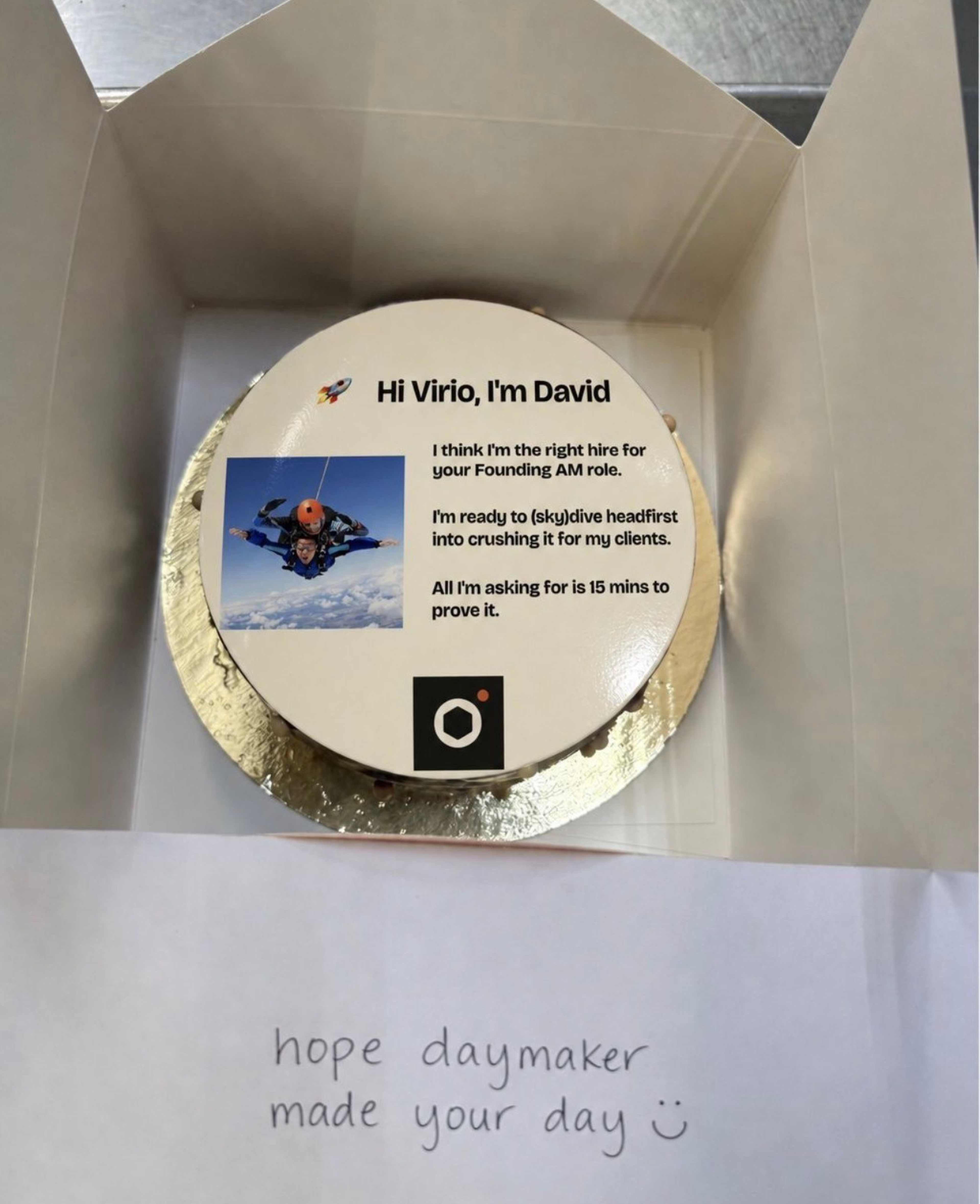 A cake with a printed photo of two people skydiving and a note from David to Virio, expressing readiness for a Founding AM role, plus a handwritten paper saying, “hope daymaker made your day :)”