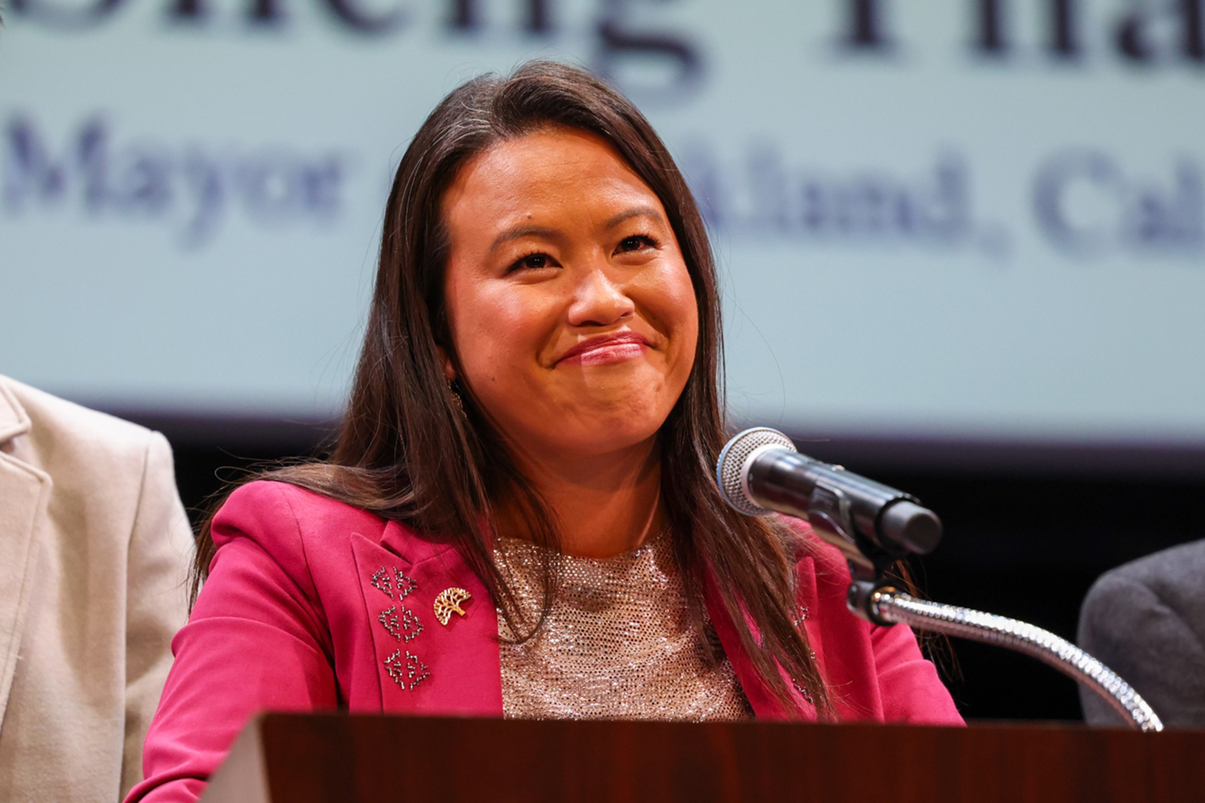 A woman in a magenta blazer and light-colored top is standing at a podium with a microphone, smiling. There is text in the blurred background.