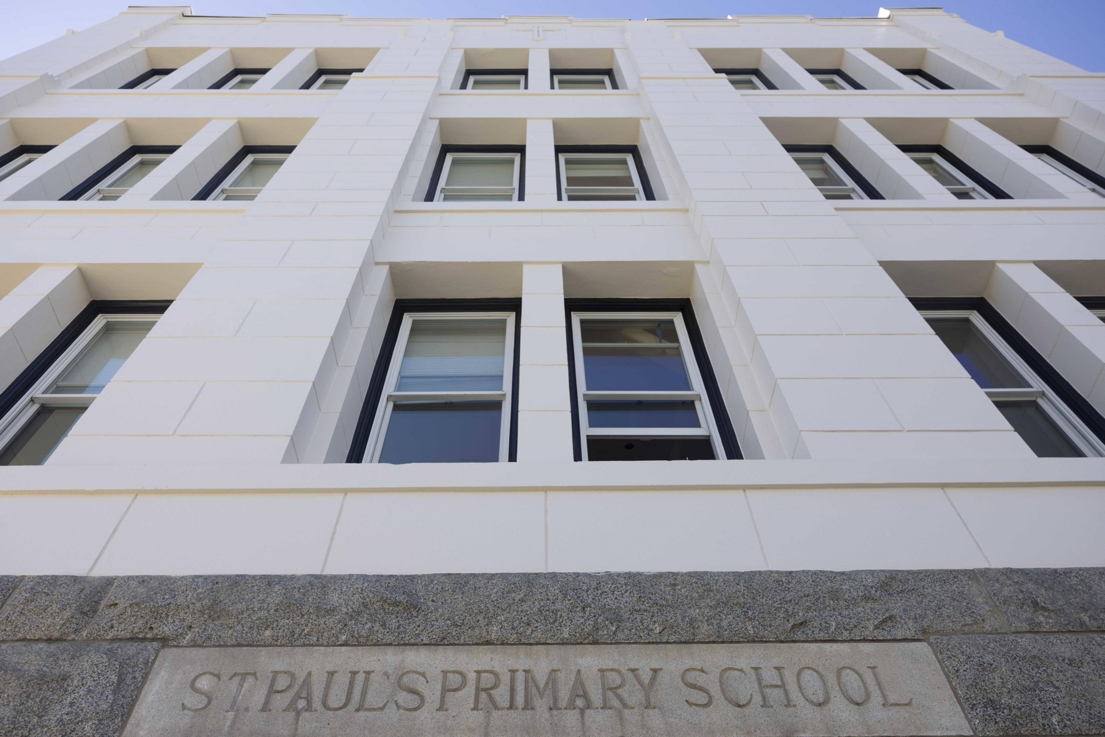 The image shows a white building with a modern design featuring large windows. The stone plaque at the base reads &quot;St. Paul's Primary School.&quot;