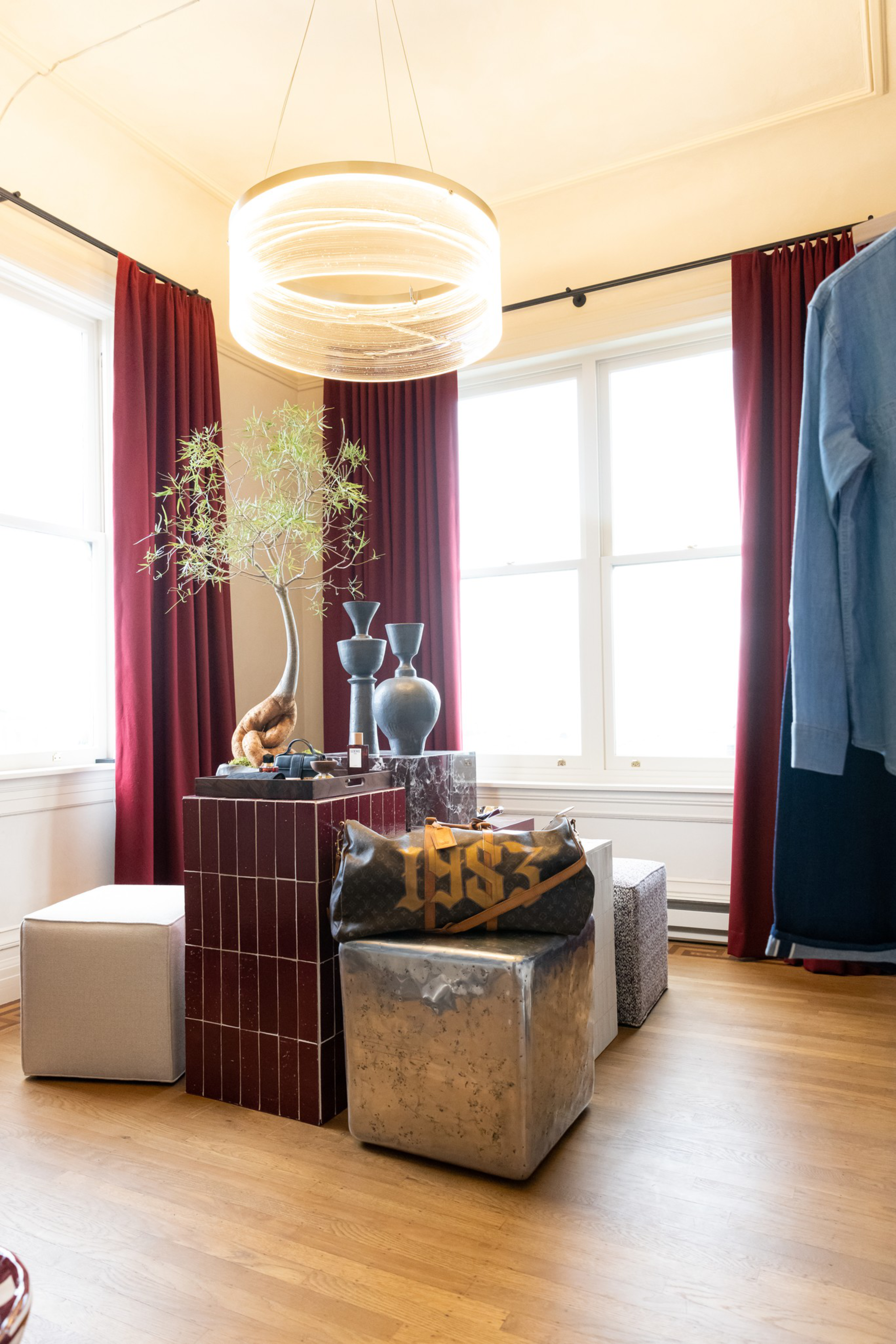 A cozy corner with burgundy curtains, a potted tree on a tiled pedestal, two vases, cube-shaped ottomans, and a duffel bag on wood flooring.