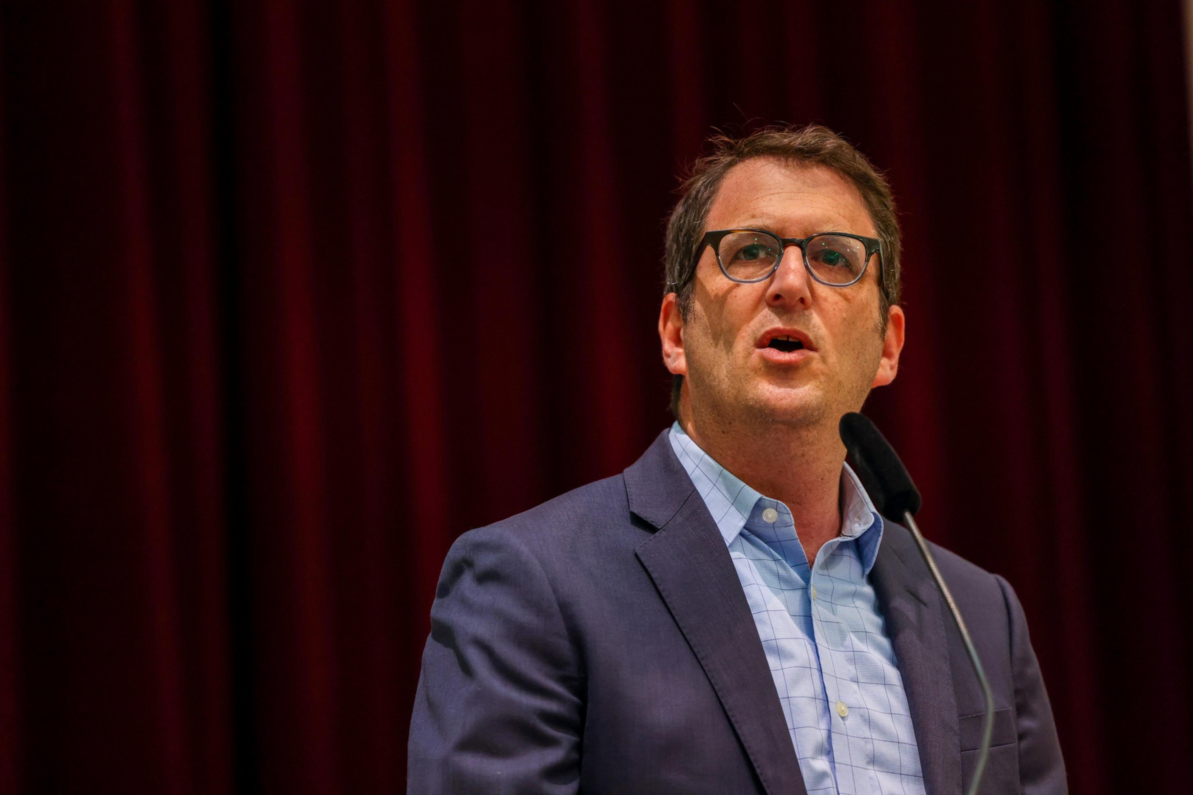 A man in glasses and business attire speaks at a podium against a red curtain backdrop.
