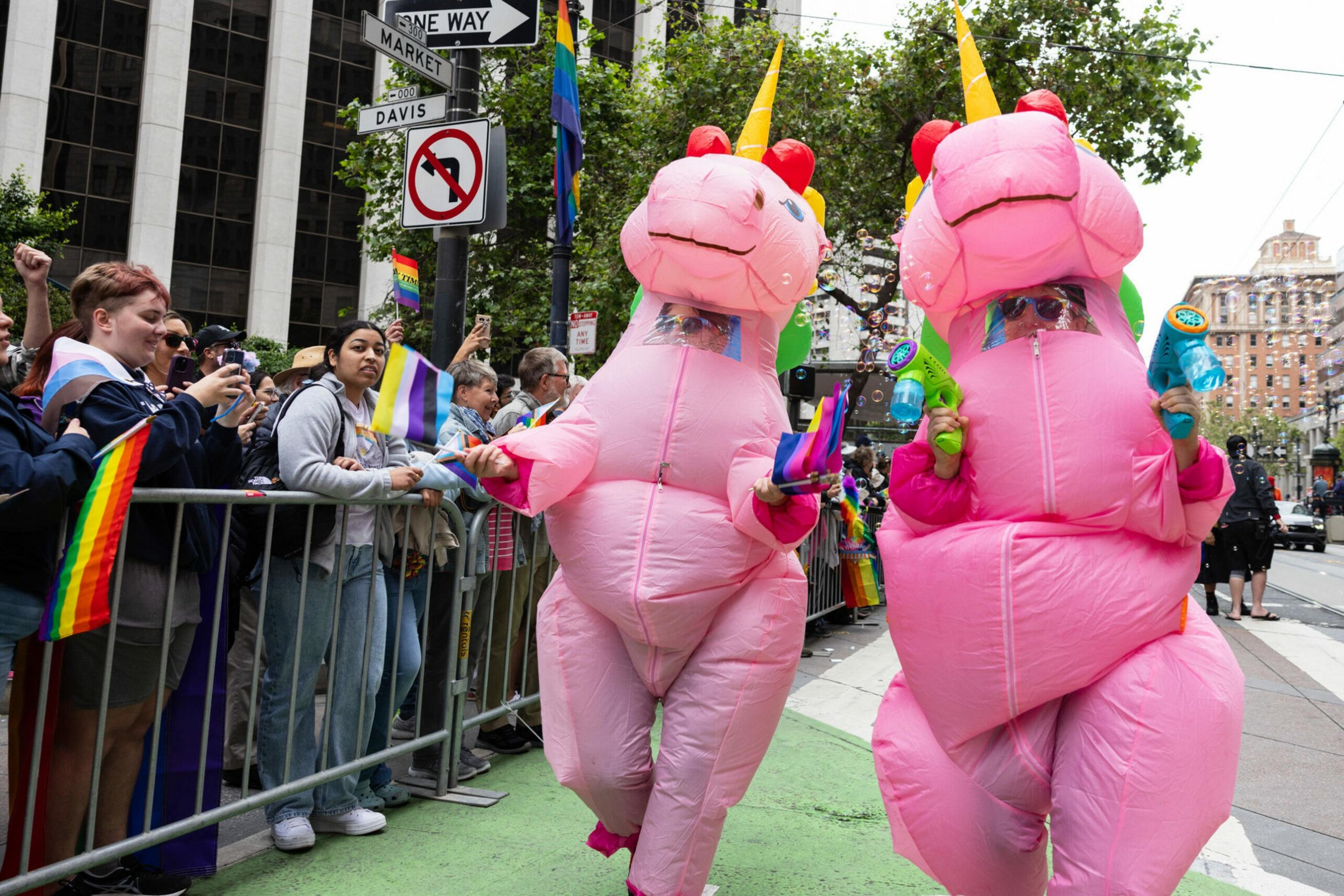 Two people in pink inflatable unicorn costumes hold colorful water guns, walking past a crowd waving rainbow and pride flags on a city street.
