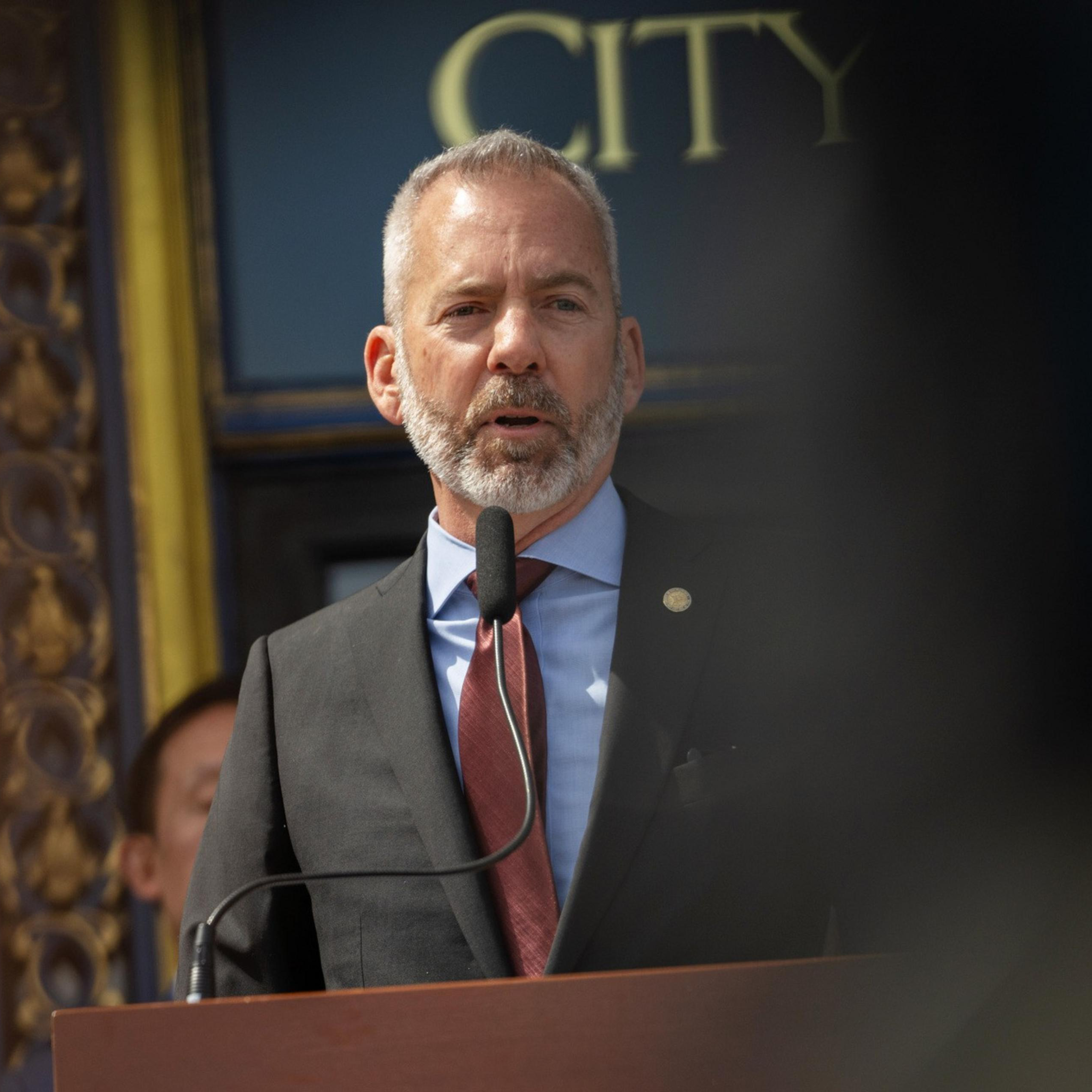 A man with a beard in a suit and red tie speaks at a podium with a microphone, in front of a decorative background. Part of the word "CITY" is visible.