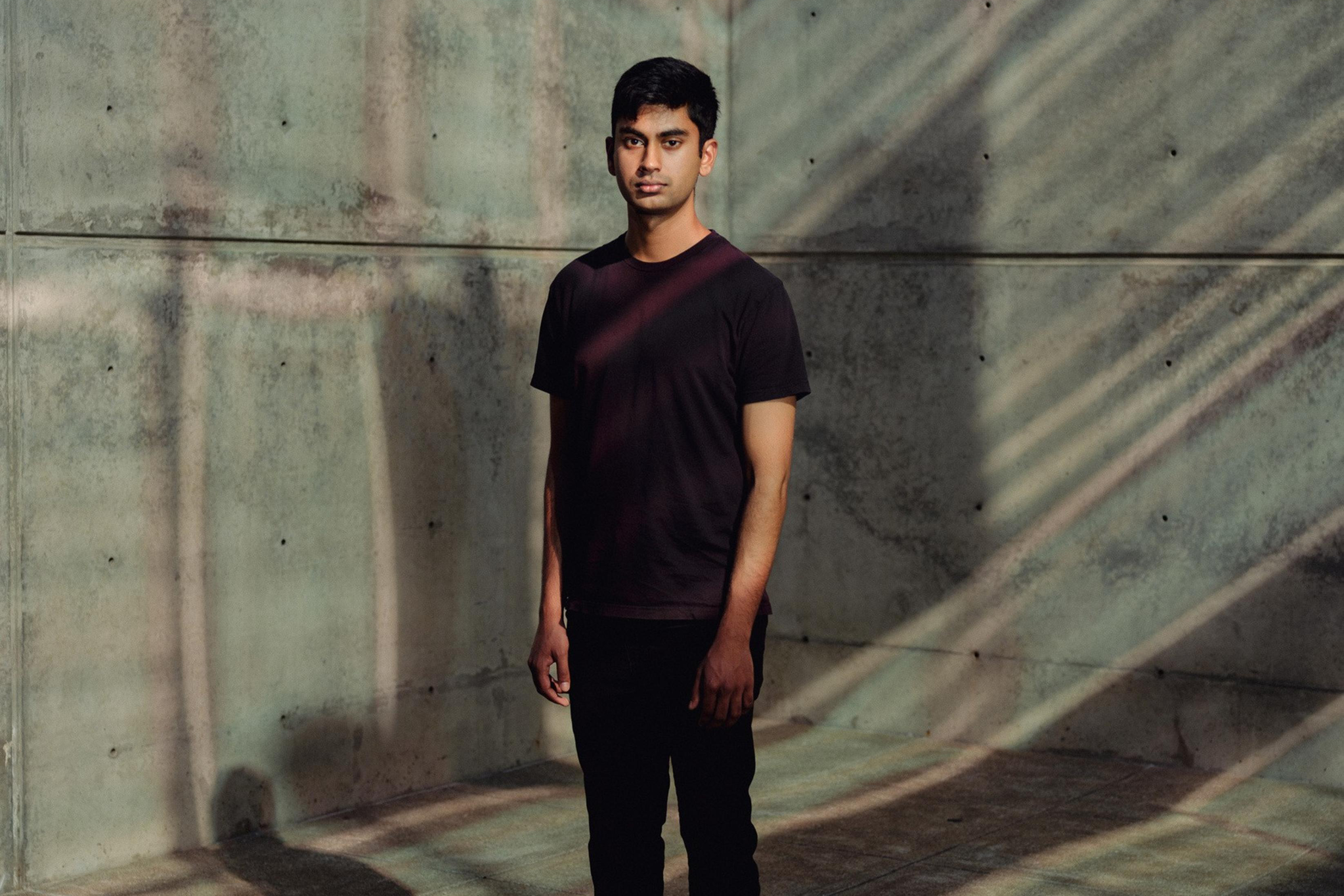 Suchir Balaji stands in a concrete-walled space, illuminated by diagonal light and shadow patterns. He wears a black shirt, dark pants, and white sneakers.