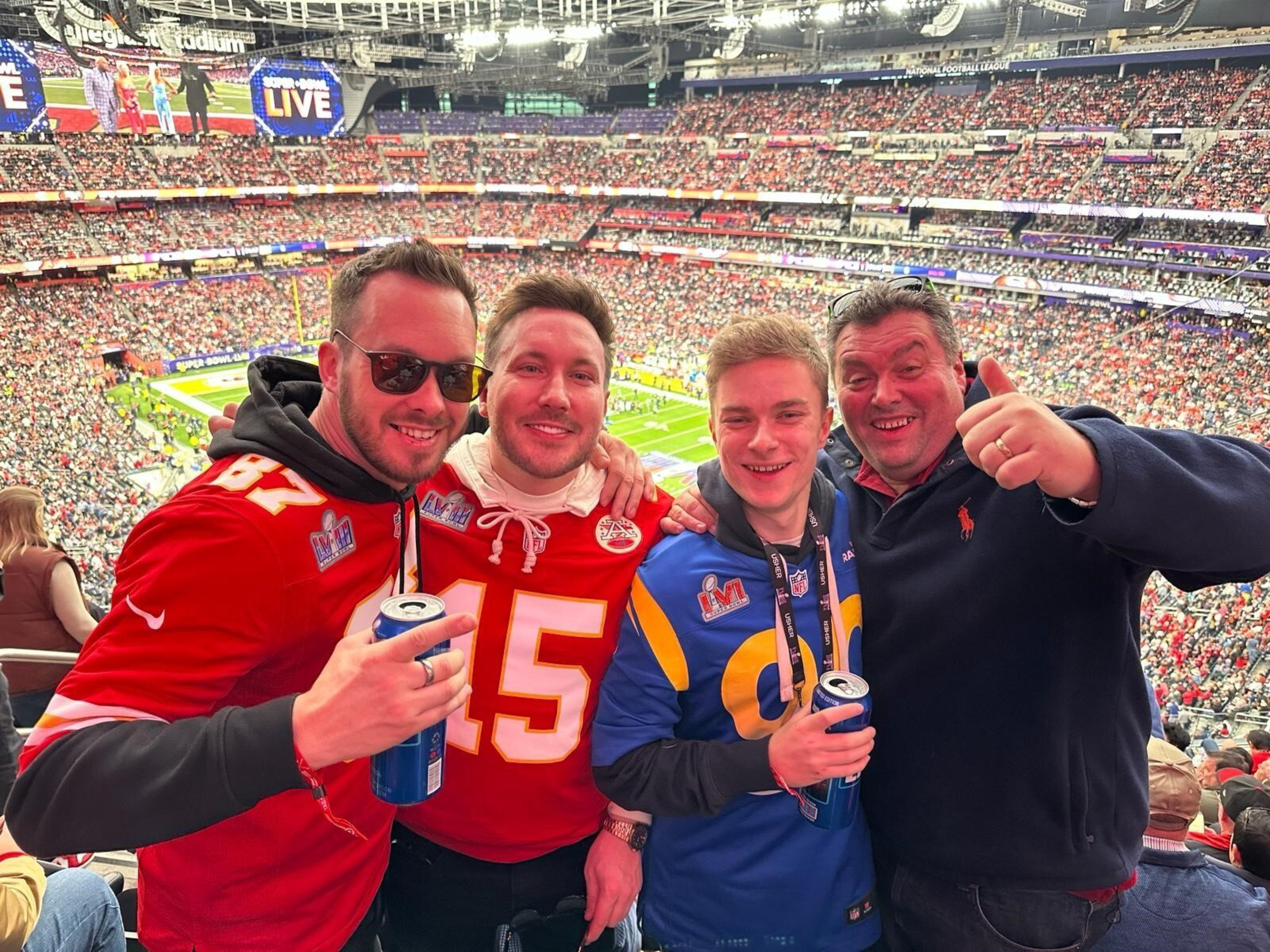 Four men smiling and posing for a photo at a packed football stadium, two wearing red Chiefs jerseys and one in a blue Rams jersey, holding drinks.