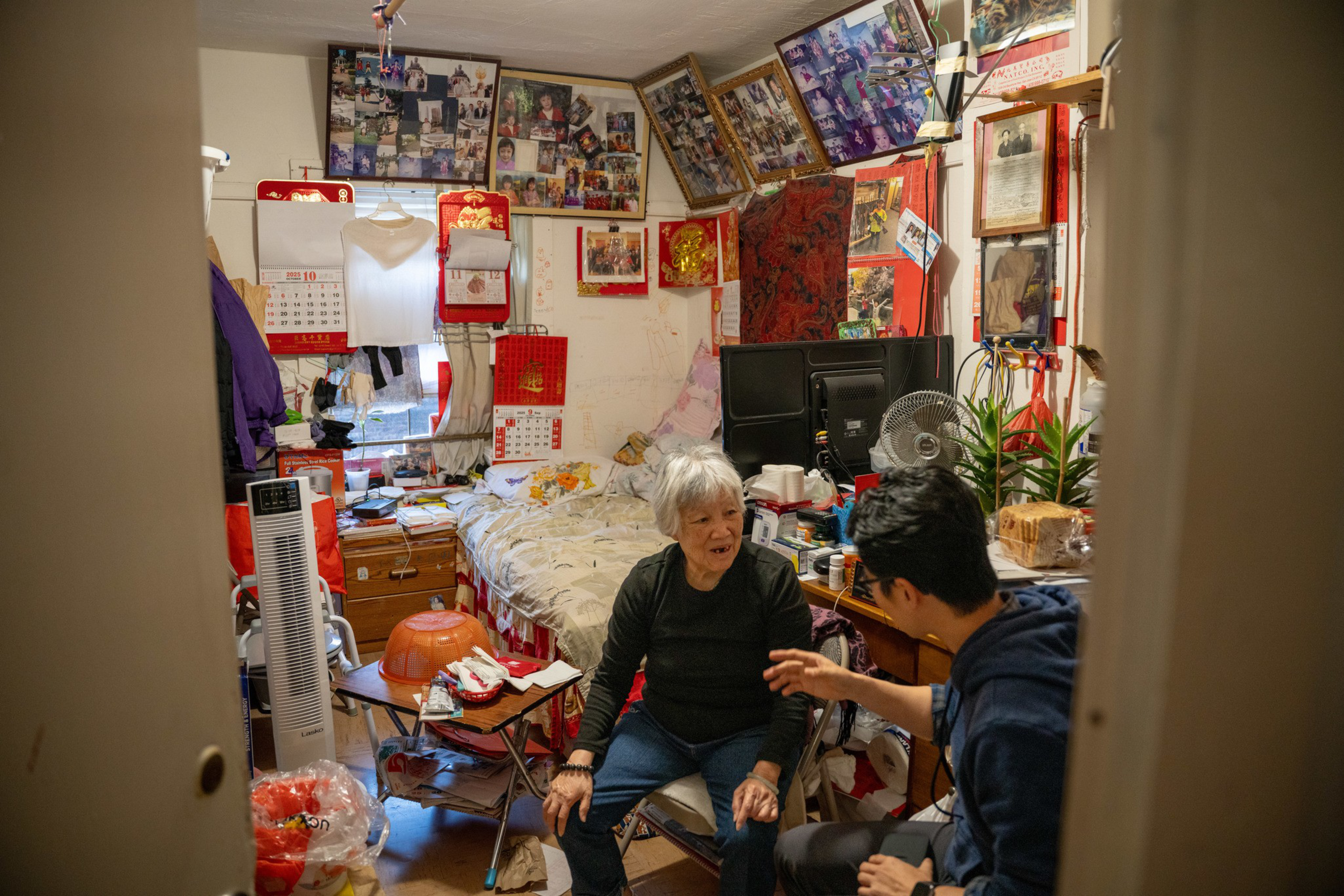 An elderly woman and a younger man sit and talk in a cluttered, small room filled with photos, calendars, a bed, and various household items.