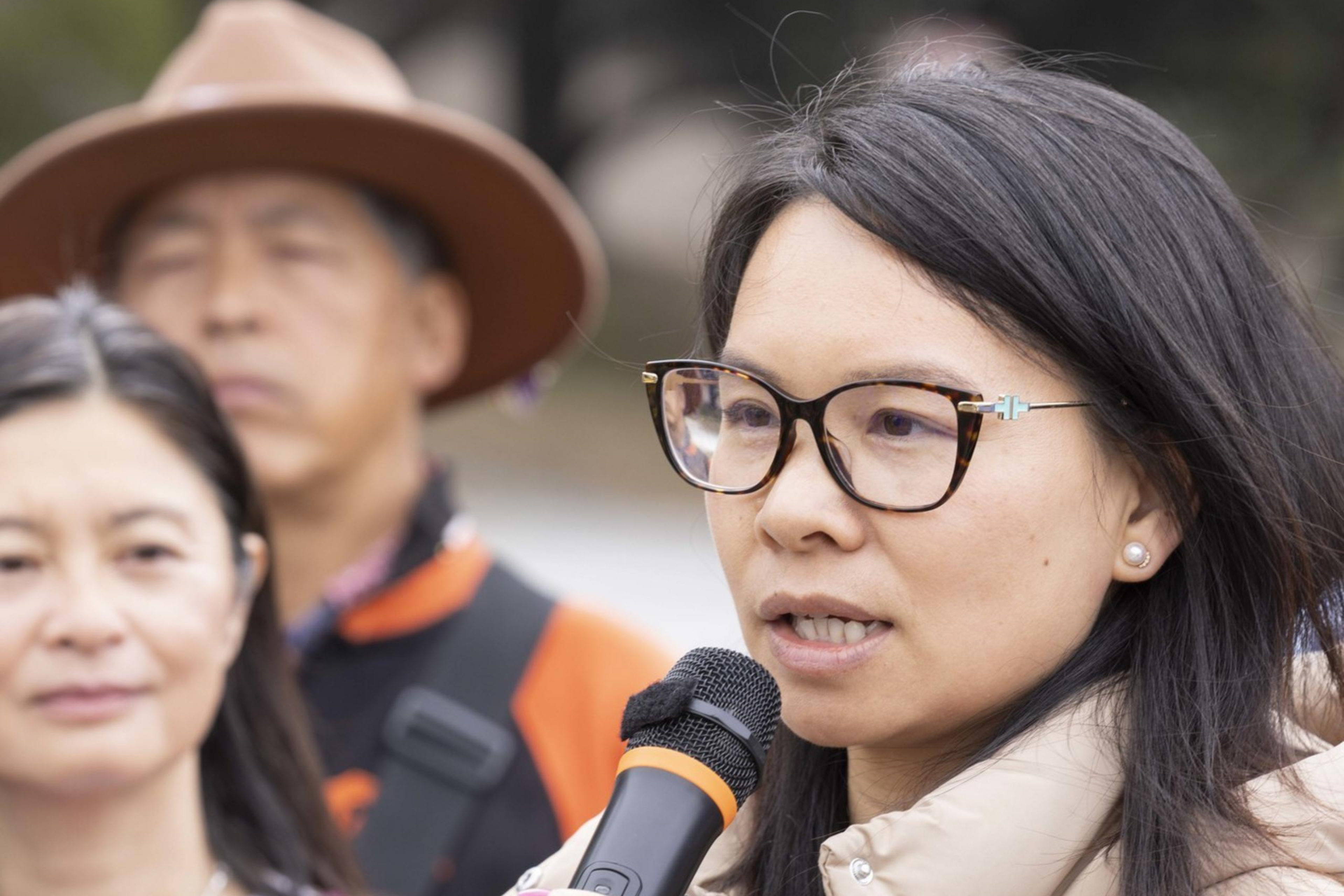 A woman in glasses speaks into a microphone, wearing a beige jacket. Behind her, another woman and a man in a hat stand, holding a blue sign with partial text.