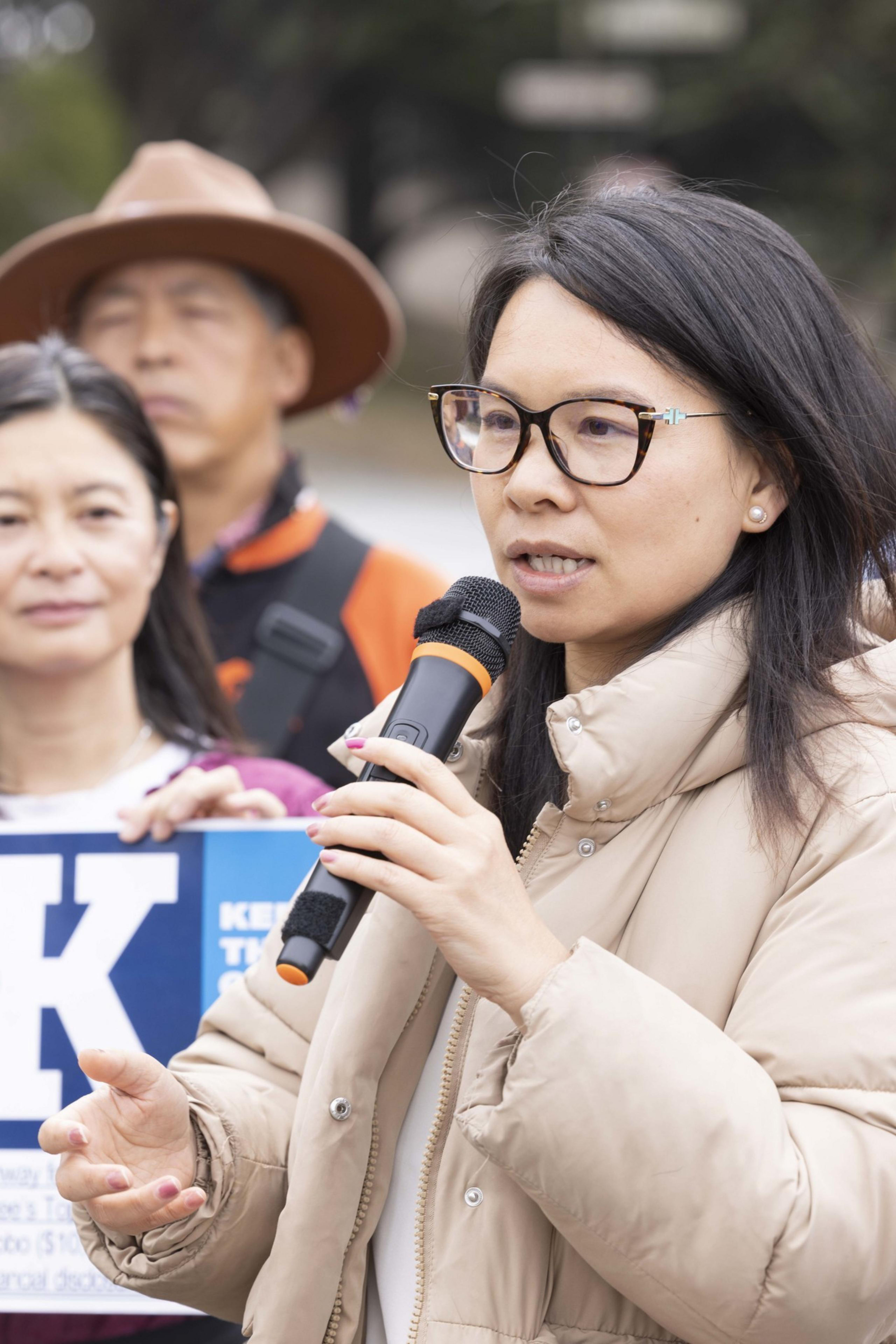 A woman in glasses speaks into a microphone, wearing a beige jacket. Behind her, another woman and a man in a hat stand, holding a blue sign with partial text.