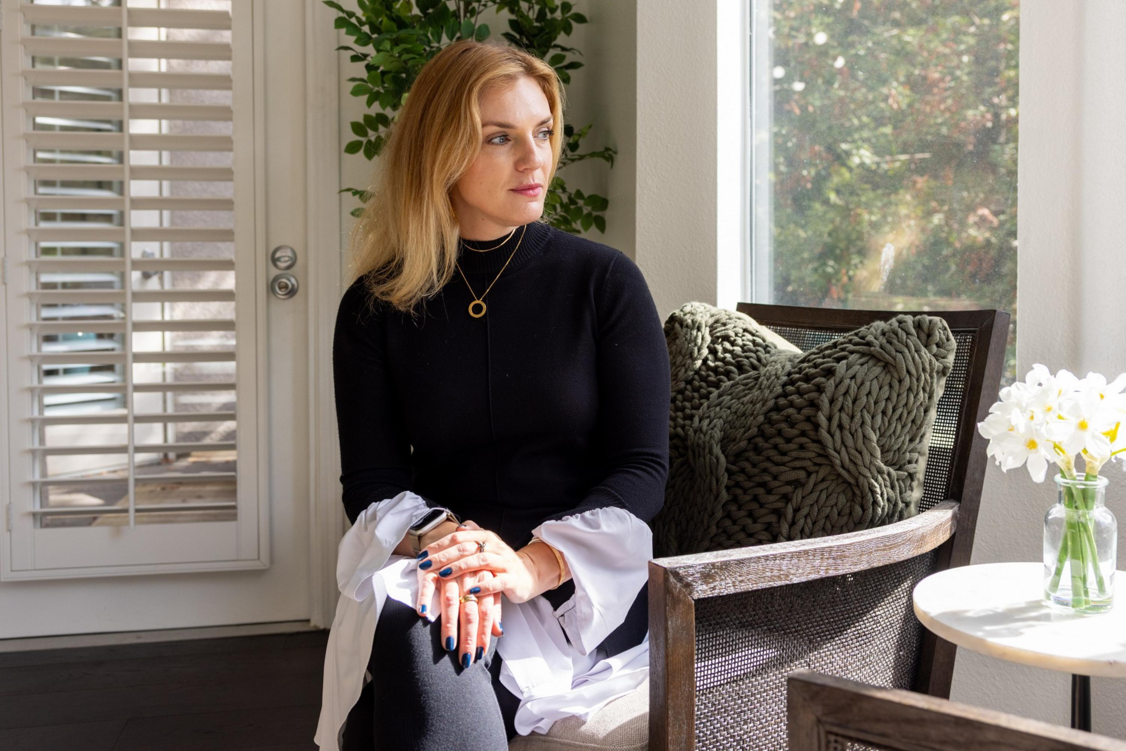 A woman with blonde hair, wearing a black top with white sleeves, sits with her hands clasped on a chair near a window with sunlight and a vase of white flowers nearby.