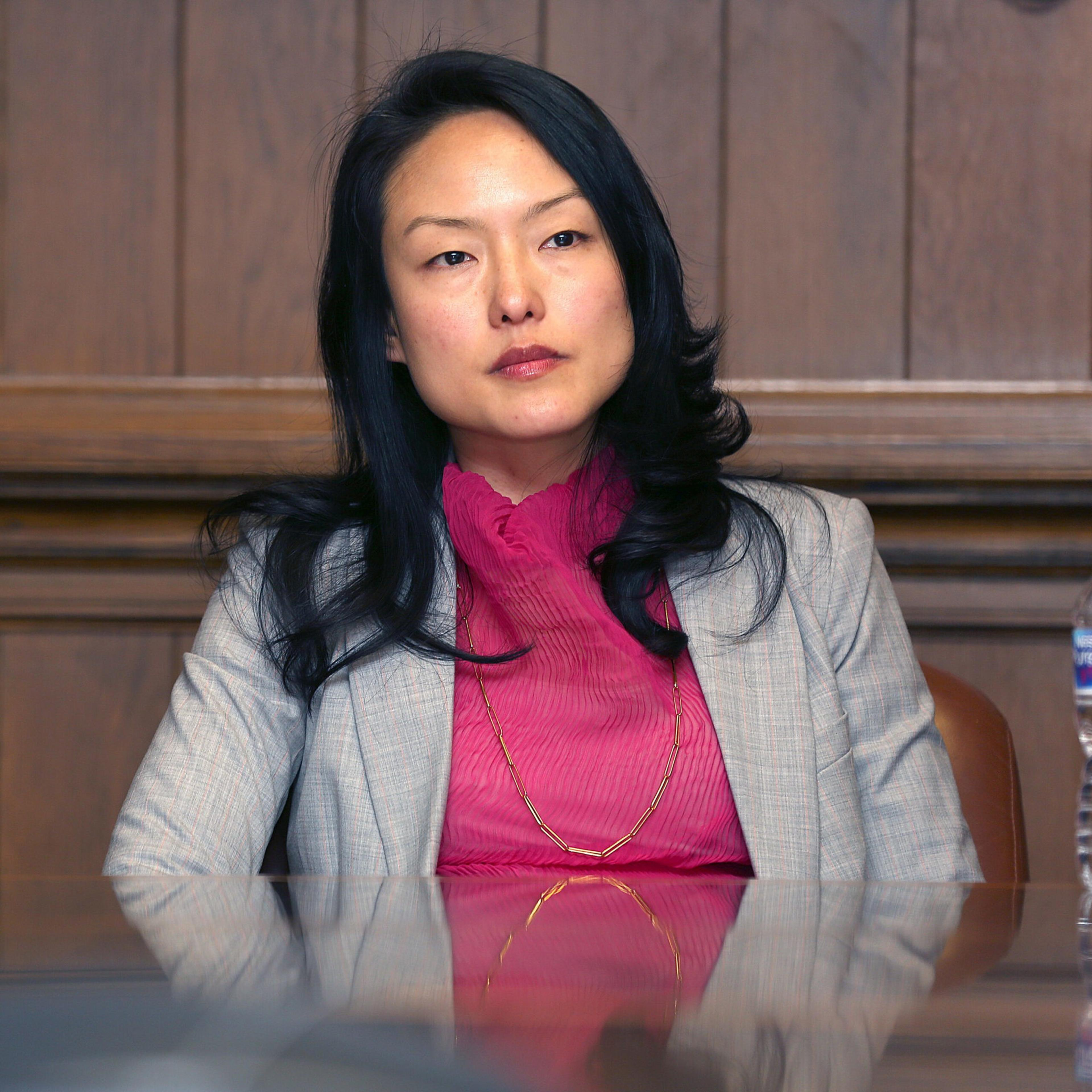 A woman with long black hair wears a pink blouse and light gray blazer, sitting at a wooden table with a water bottle to her right.
