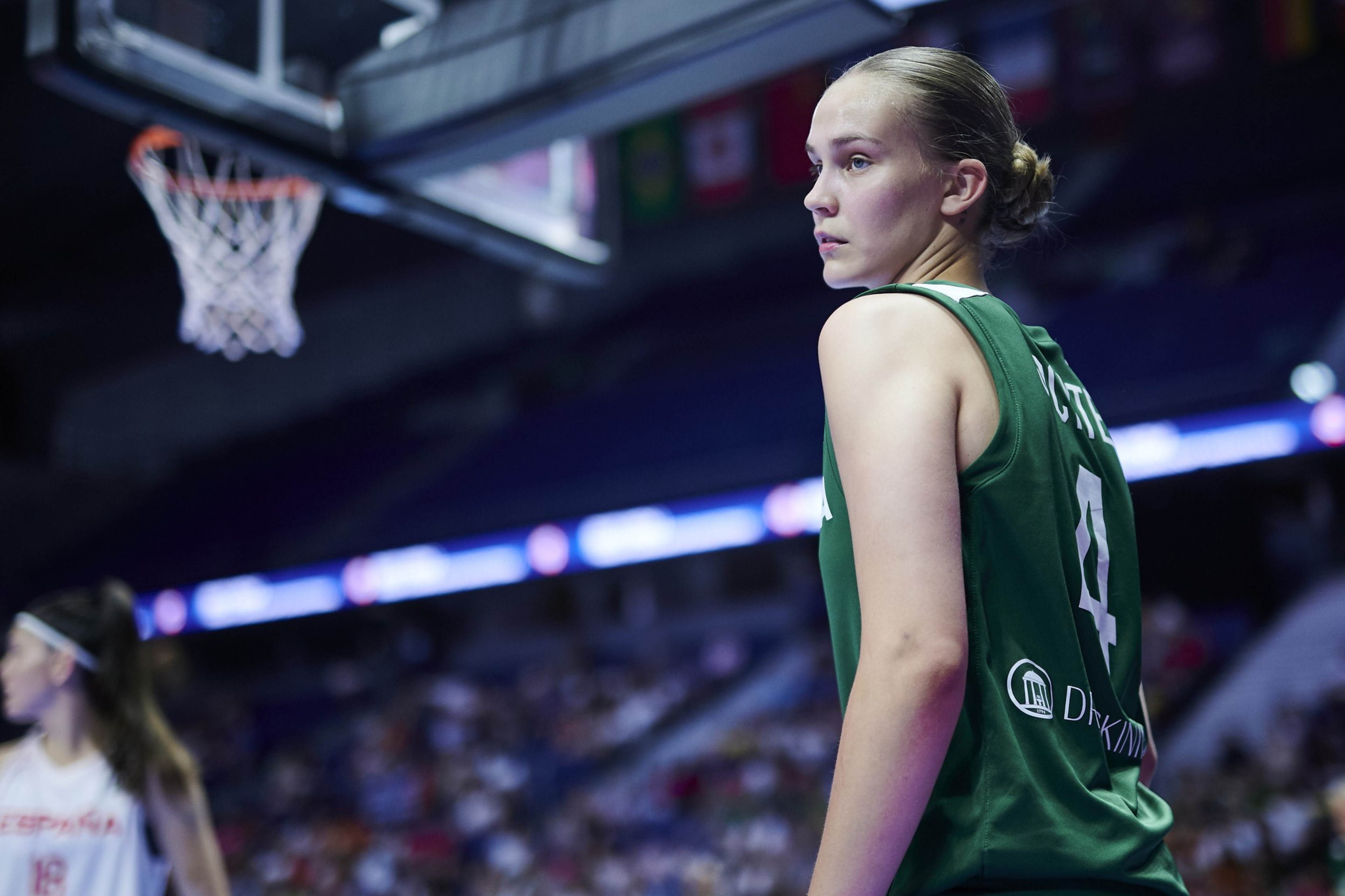 A basketball player in a green jersey looks over her shoulder on the court, with a blurred basketball hoop above and a crowded stadium in the background.