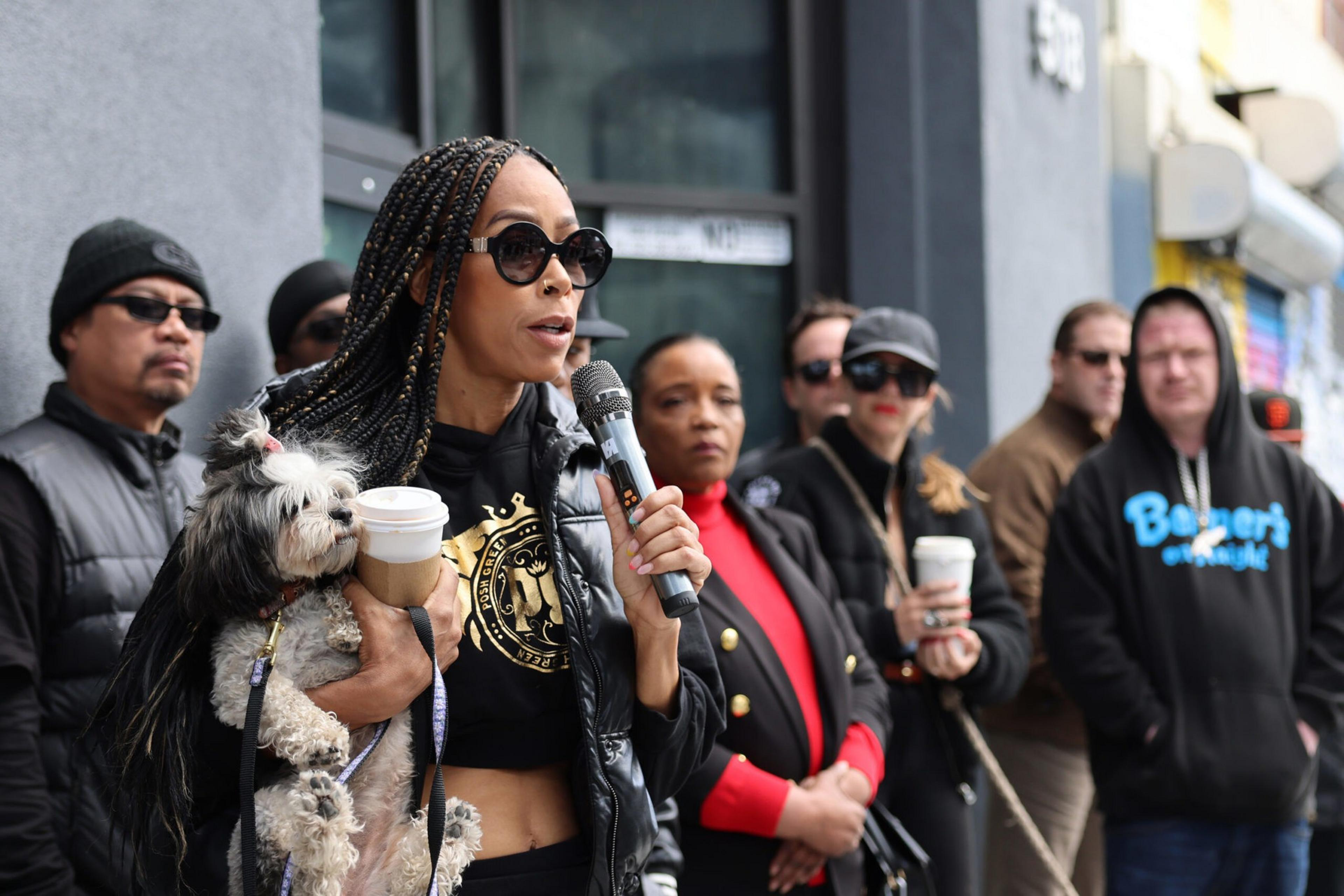 A woman with braided hair and sunglasses holds a small dog and coffee while speaking into a microphone, with a group of people standing behind her.
