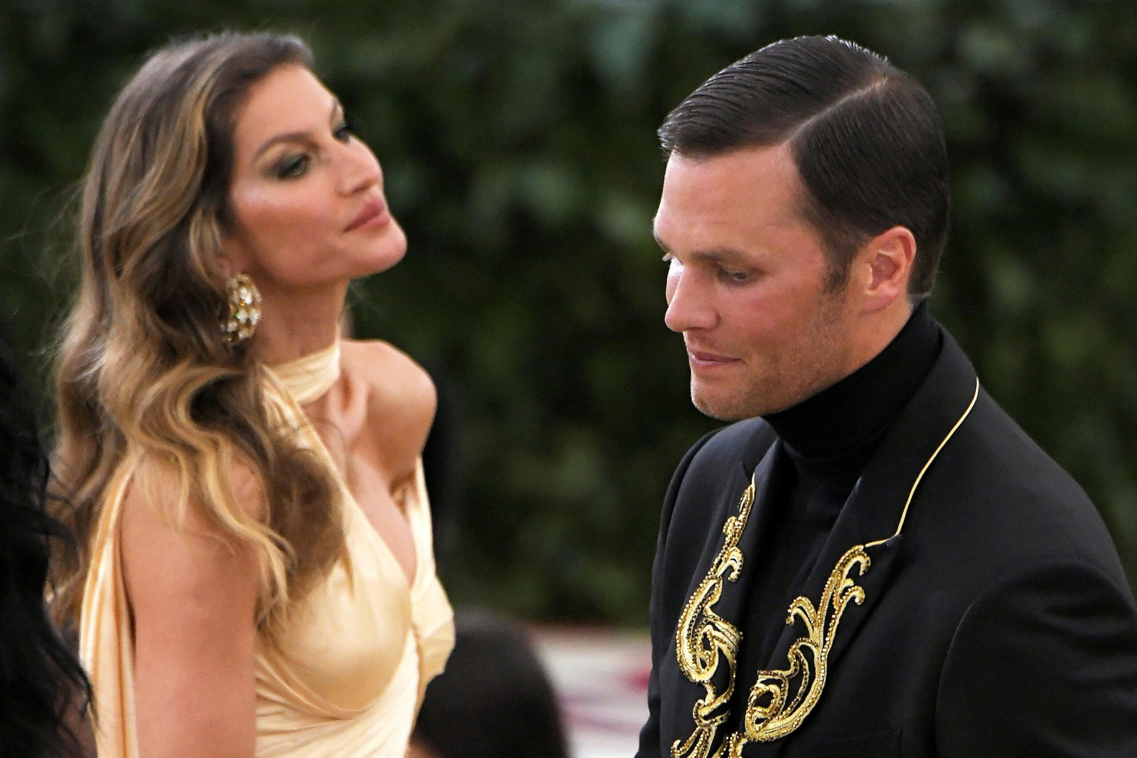 A woman in a light gold dress with large earrings looks at a man in a black jacket with gold embroidery, who is looking slightly downward.