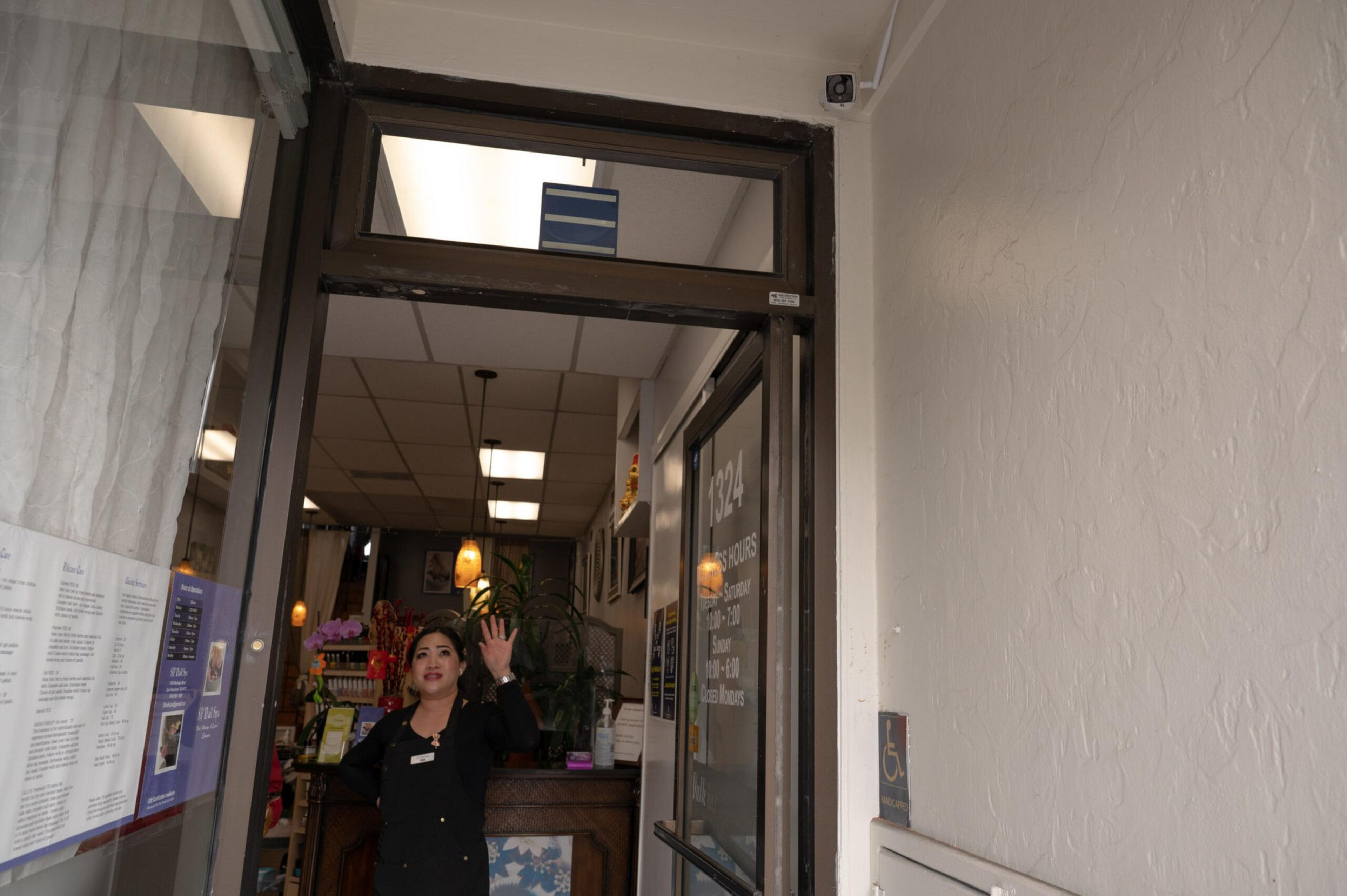 A woman in a black uniform waves from inside a well-lit storefront with plants, hanging lights, and menus posted near the entrance.