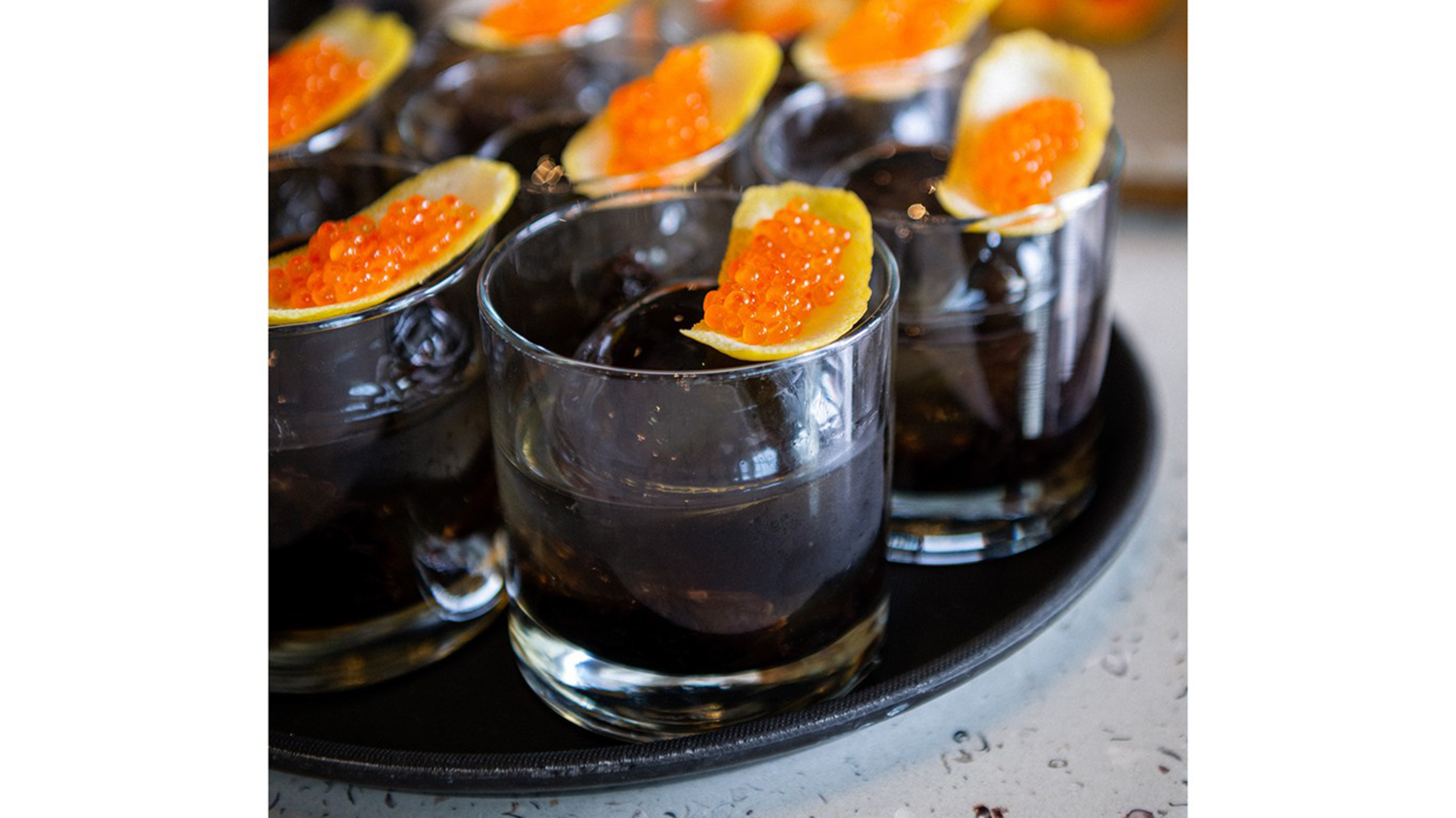 Clear glasses filled with a dark liquid and ice are each topped with a slice of fruit holding bright orange fish roe, arranged on a black tray.