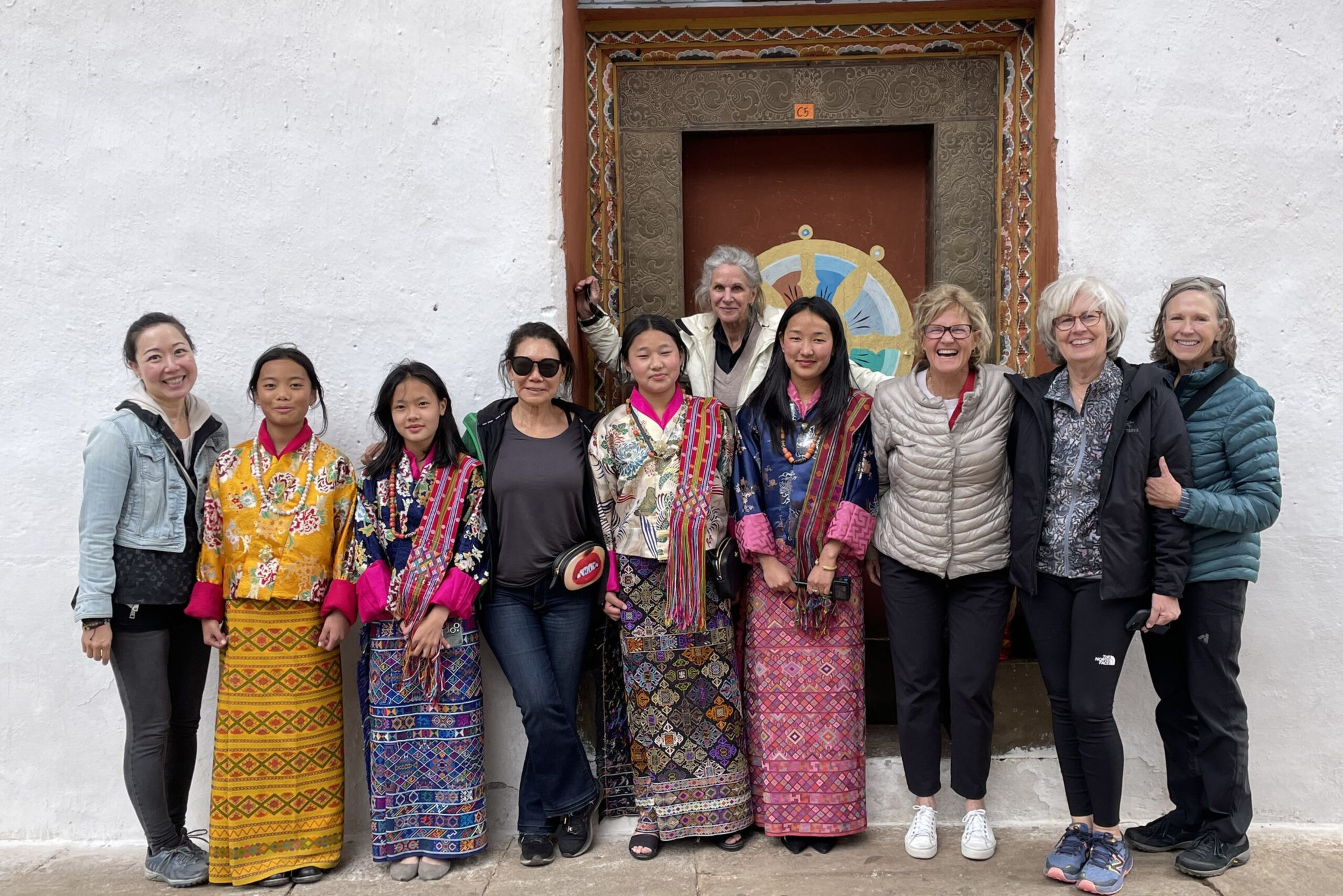 Nine women stand in front of a decorated door; four wear colorful traditional dresses, while five wear casual modern clothing.