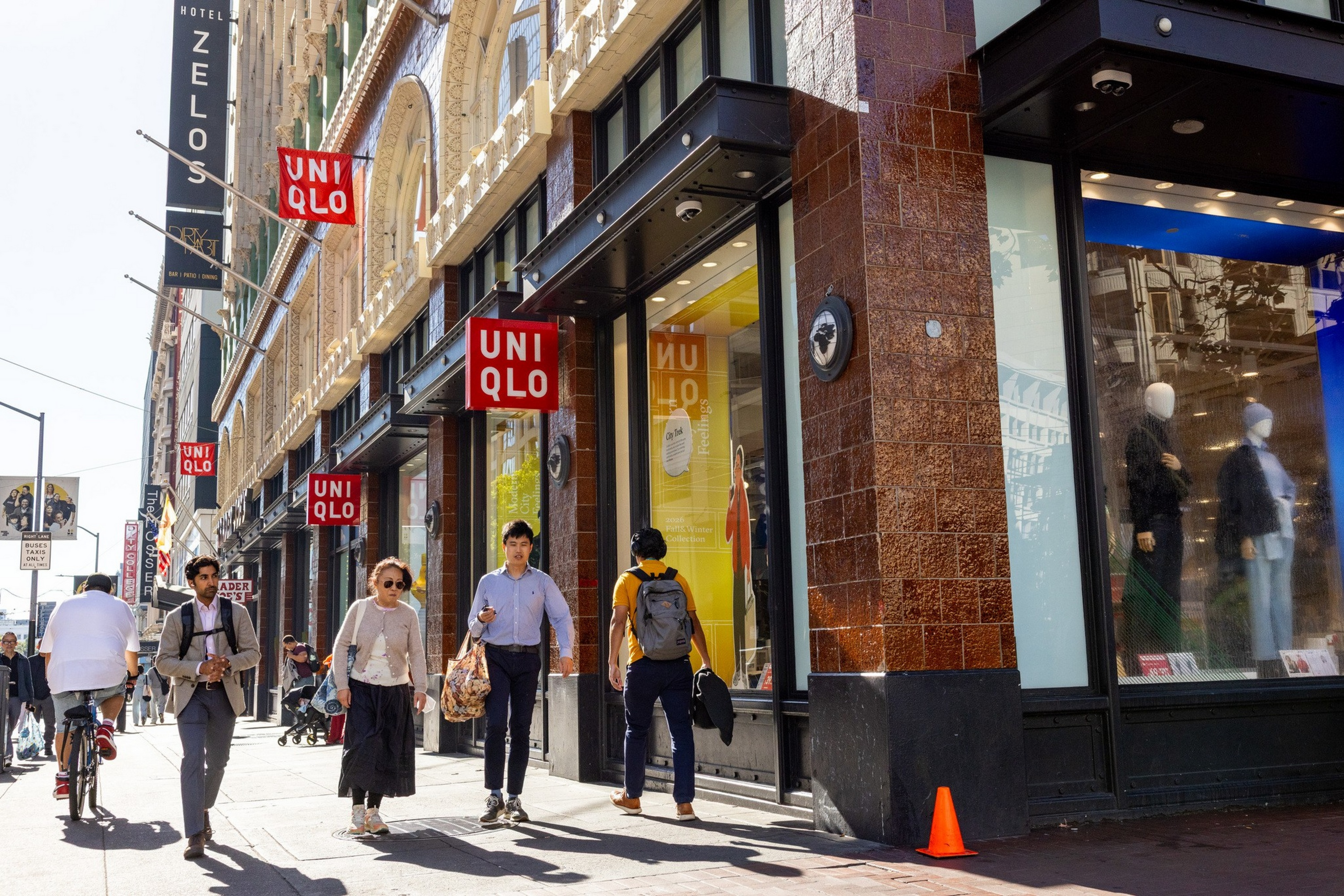 People walk past a Uniqlo store on a sunny city sidewalk, with mannequins and bright displays visible through large windows.