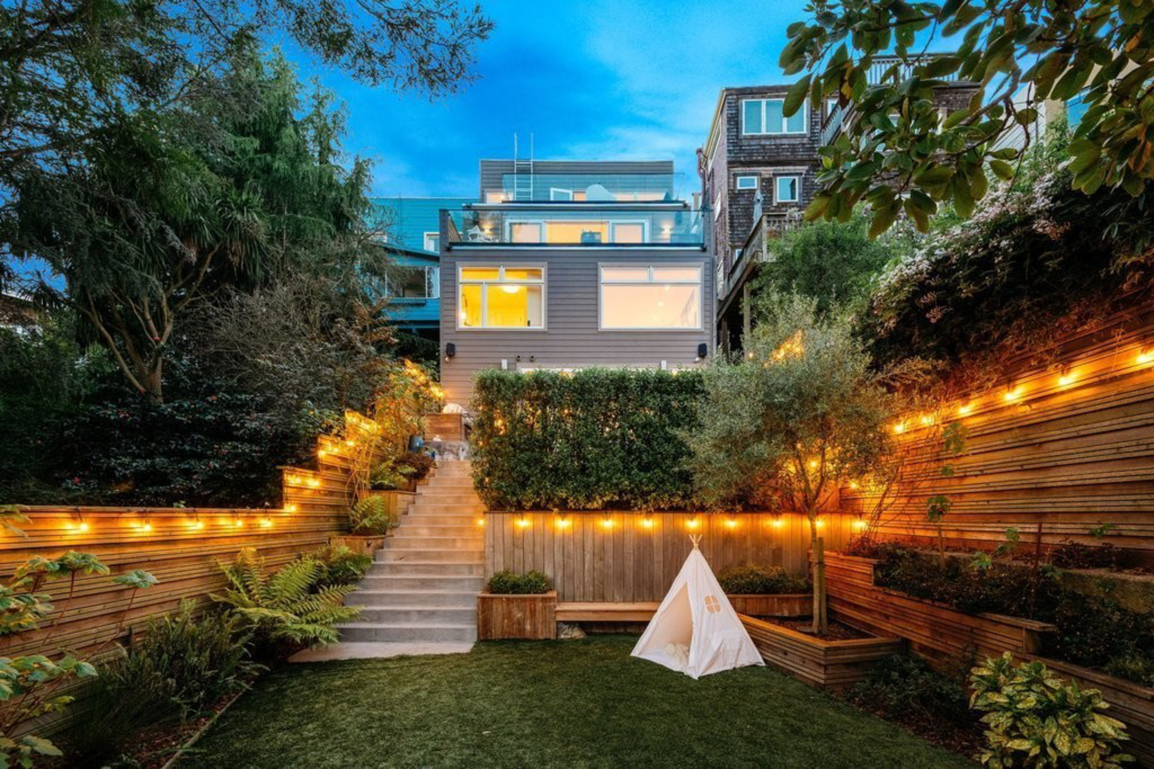 A modern backyard with string lights along wooden fences, a small white teepee on the lawn, stairs leading to a multi-story house with glowing windows.