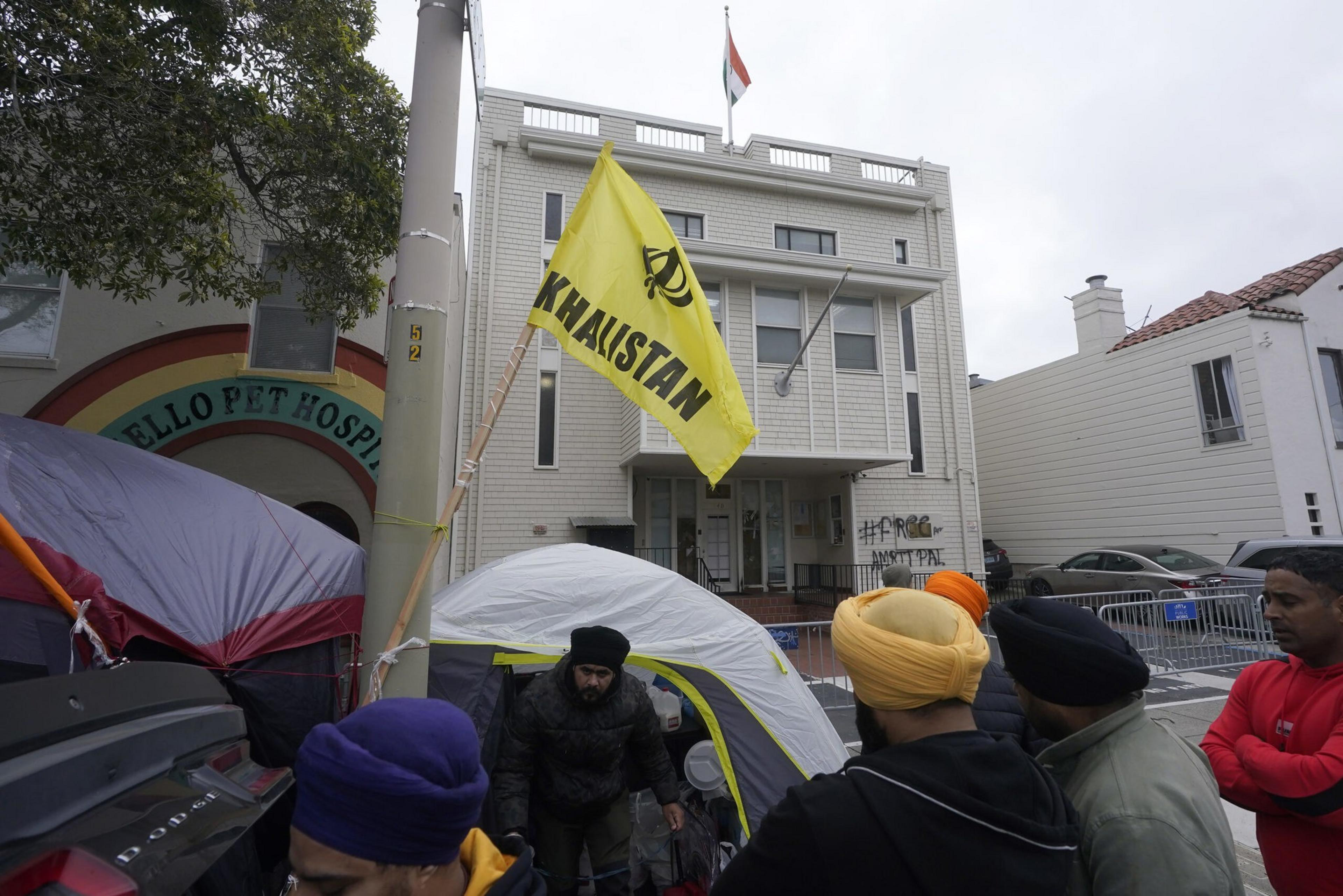 A yellow flag with "KHALISTAN" flies near tents and a group of people wearing turbans and jackets outside a white building.