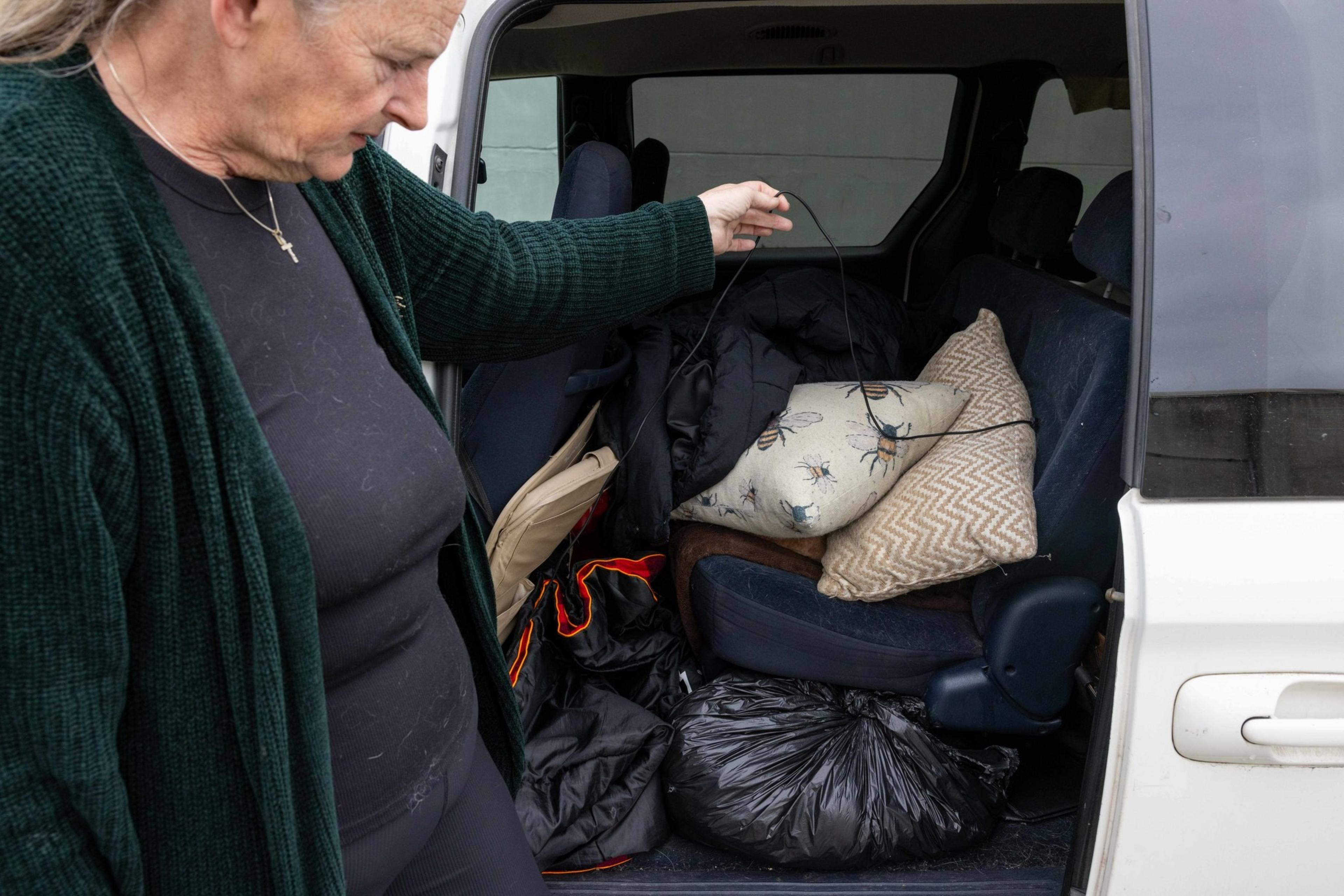 A woman stands beside a car with open doors, revealing various items inside, including bags, a jacket, and pillows with bee designs and zigzag patterns.