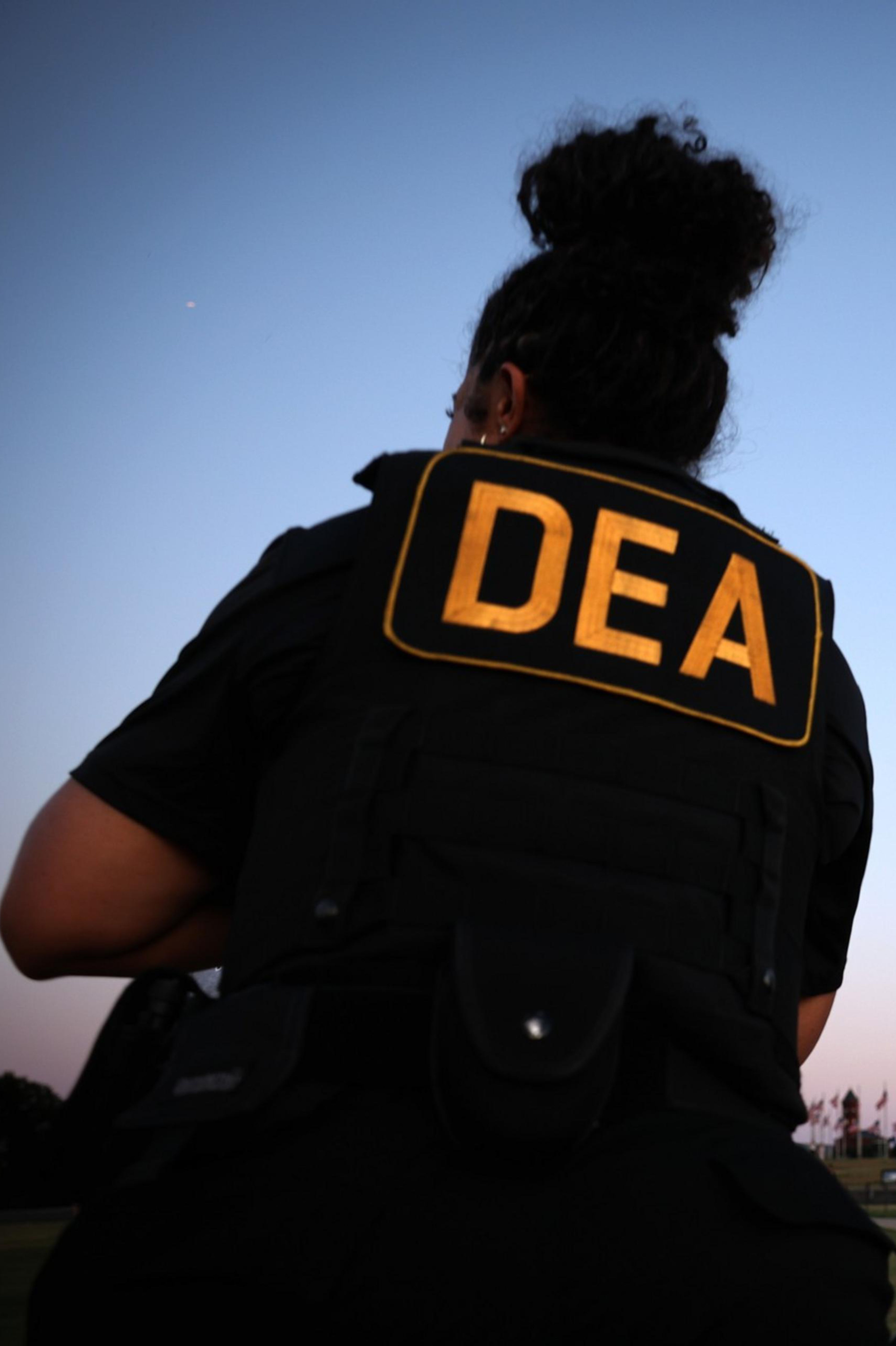 Two DEA agents stand near the Washington Monument at dusk, wearing tactical gear with “DEA” patches visible on their backs and vests.