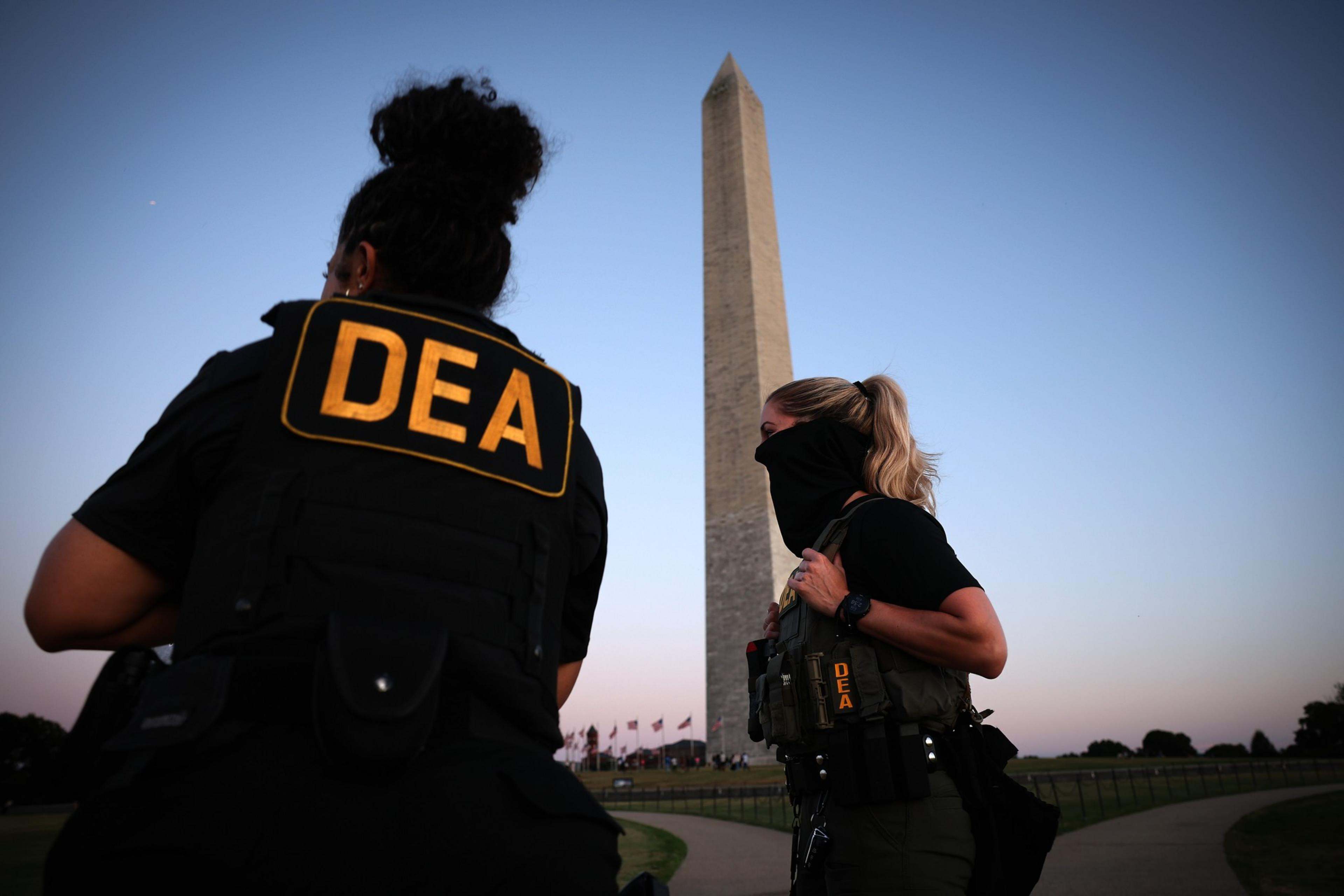 Two DEA agents stand near the Washington Monument at dusk, wearing tactical gear with “DEA” patches visible on their backs and vests.