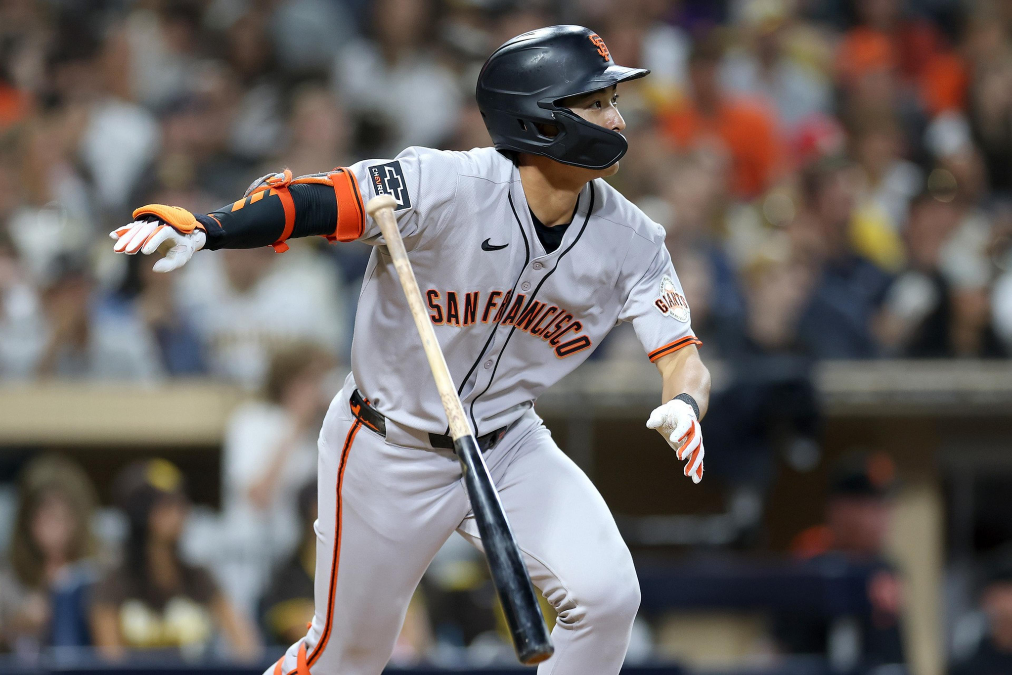 A San Francisco Giants player in gray uniform and helmet is swinging a bat during a baseball game with a blurred crowd in the background.