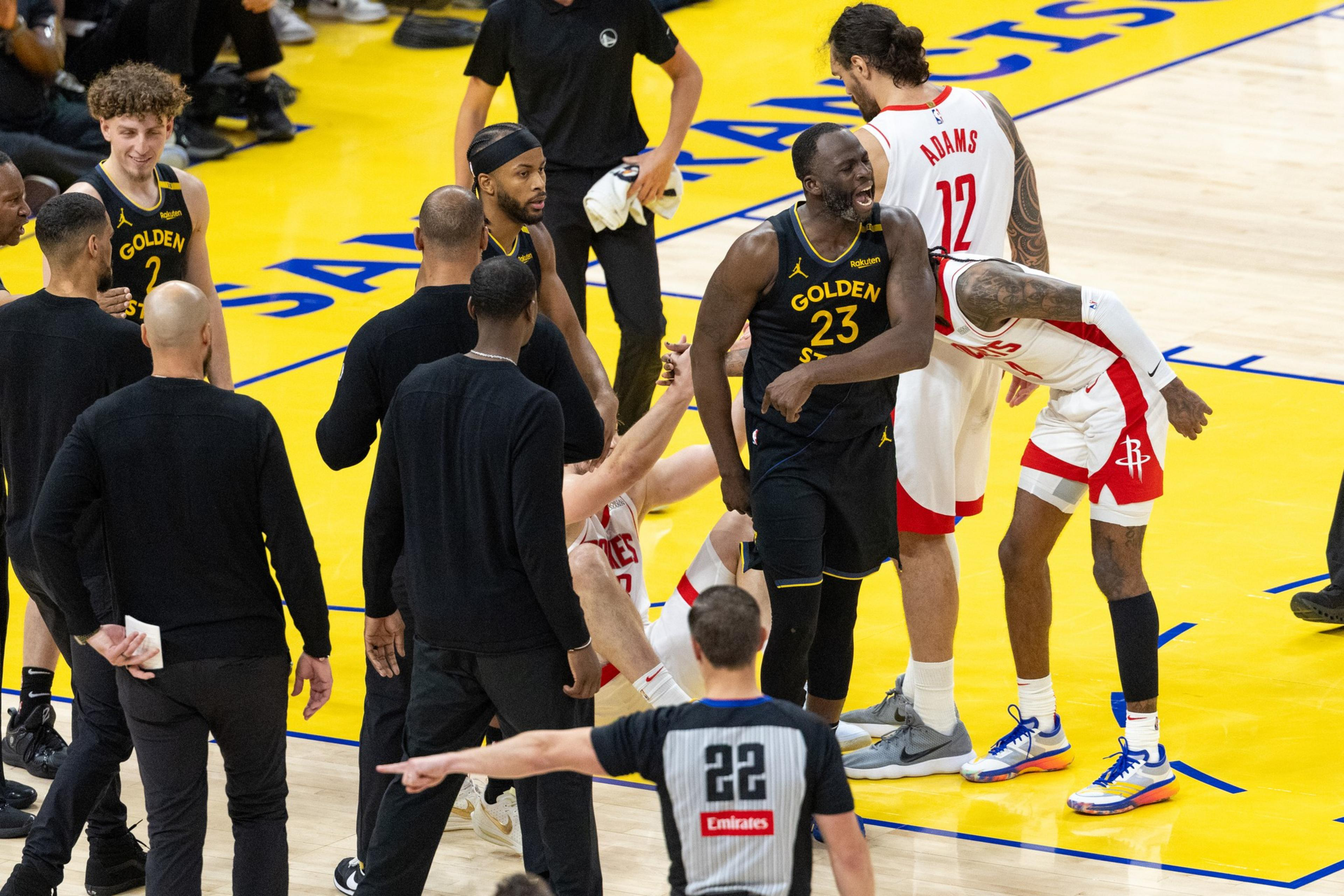 A basketball game shows players from Golden State and Houston interacting energetically on the court. One player is on the floor while others stand around him.