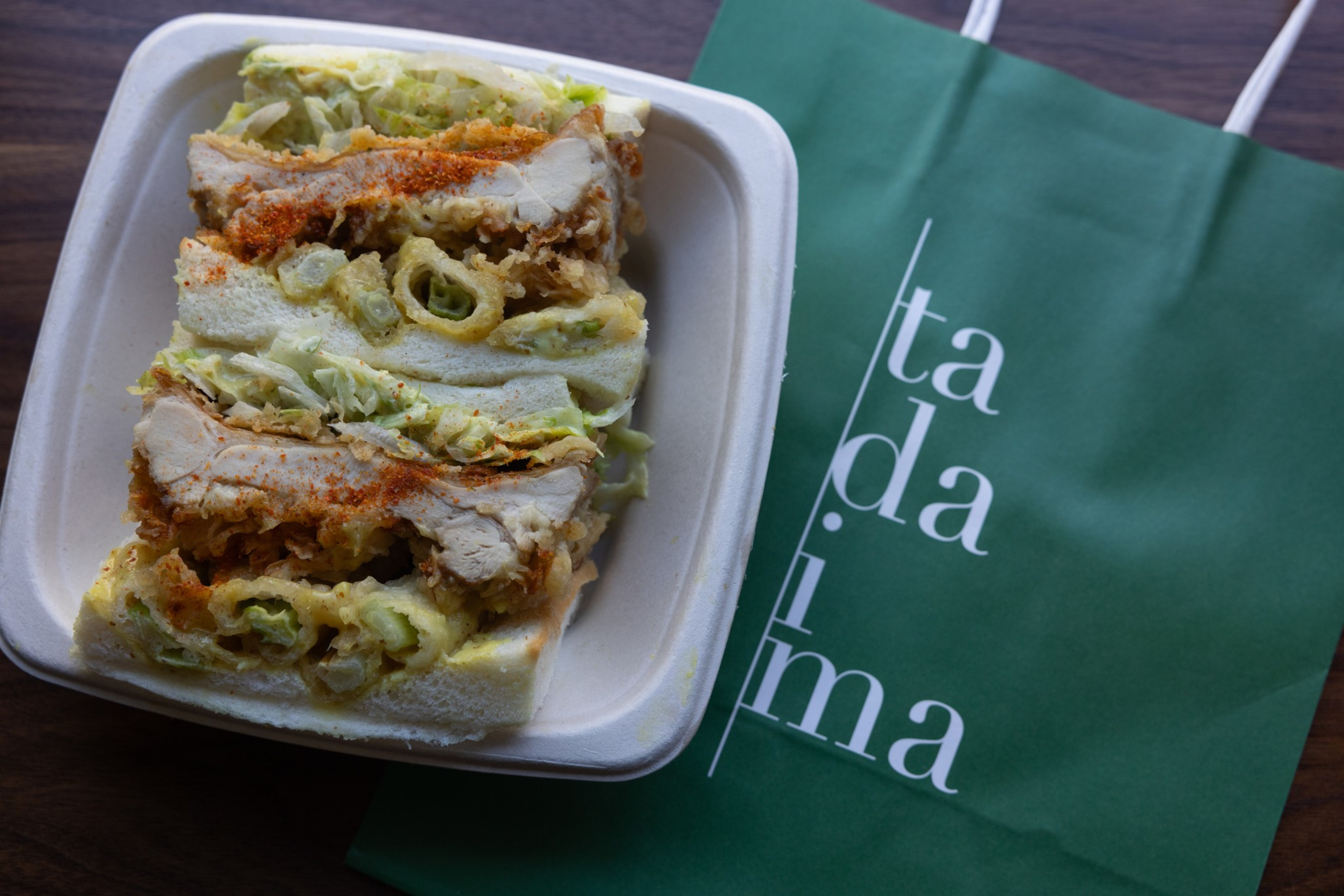 A takeout container with a chicken karage sandwich. A green bag with "tadaima" text is beside it.