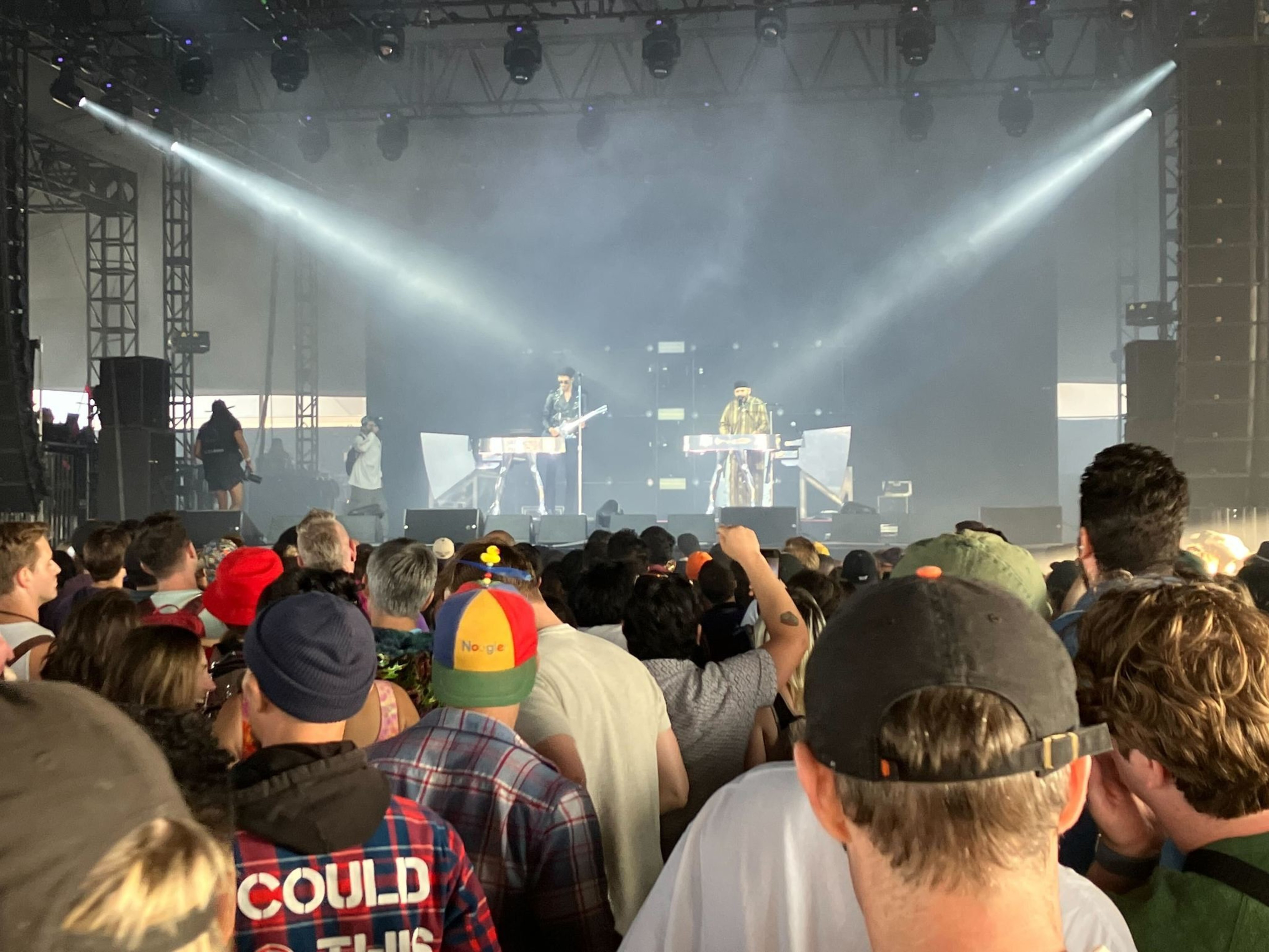A crowd watches a live band on stage with two performers playing keyboards. Bright lights shine towards the audience, creating an energetic atmosphere.
