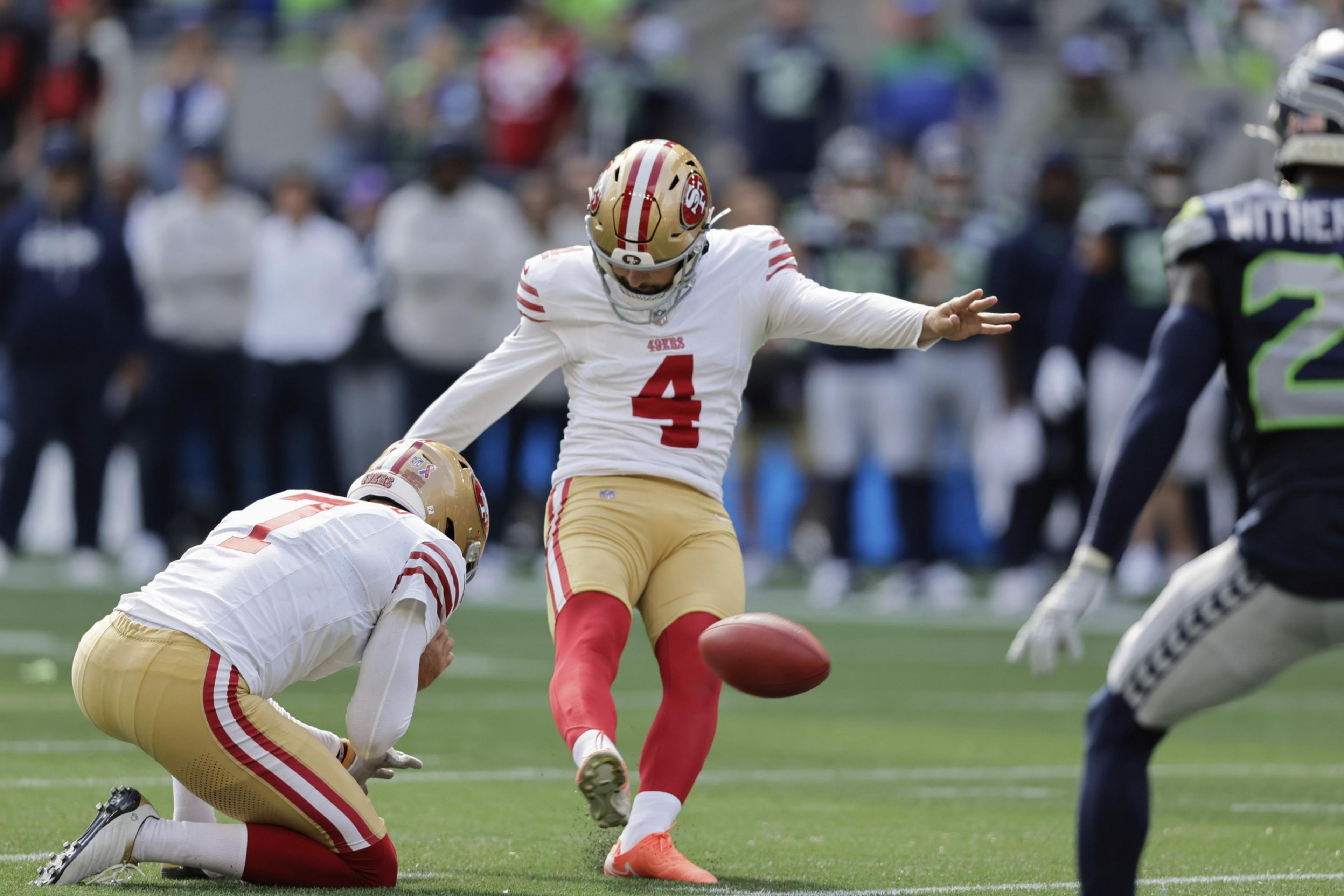 A football player wearing a white and red jersey with number 4 is kicking the ball while another player kneels holding it.