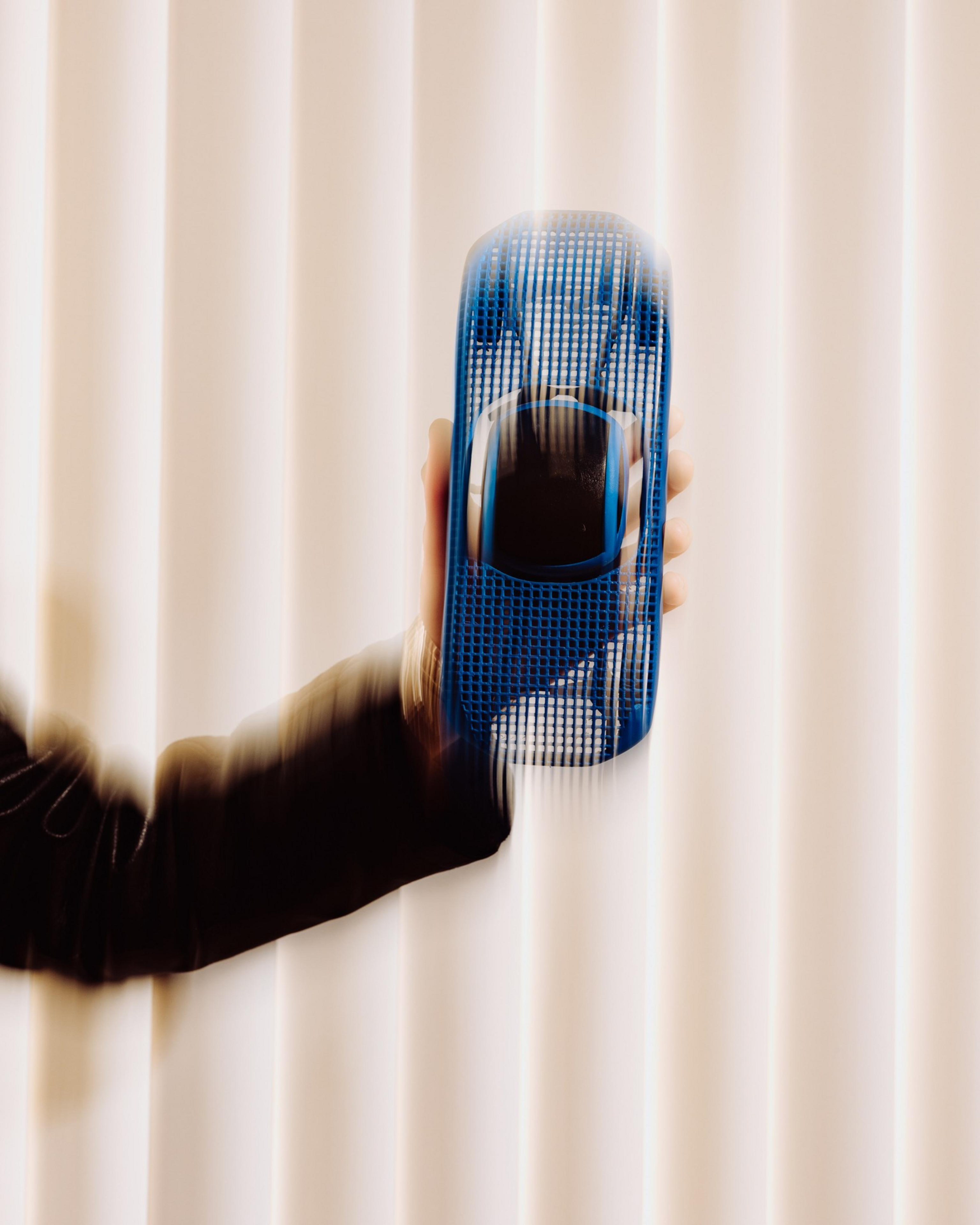 A blurred arm holds a motion-captured blue toy car against a background of vertical, light-colored blinds.