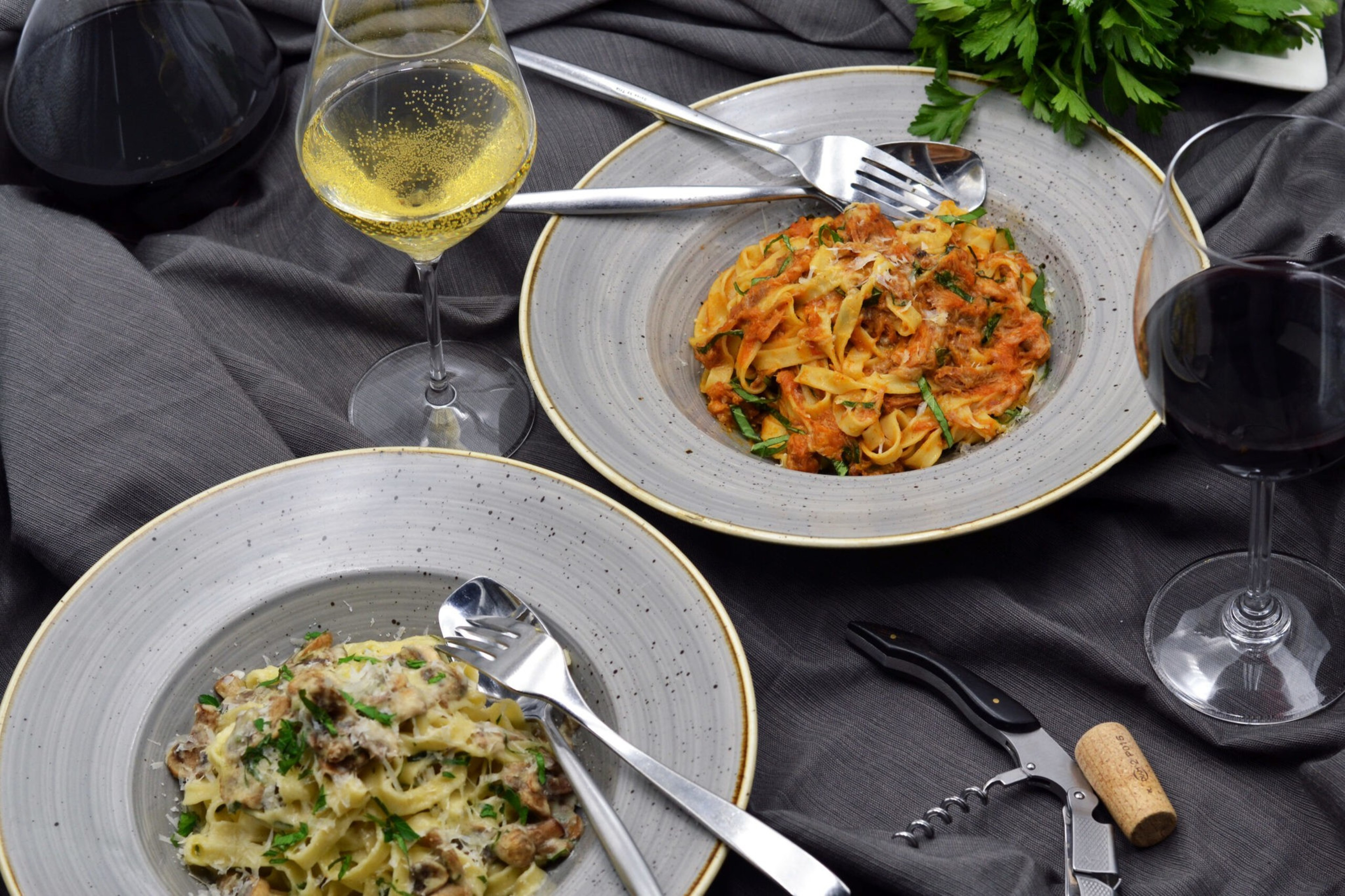 Two plates of pasta, one creamy with mushrooms and herbs, the other with a red sauce and greens, alongside glasses of white and red wine on a dark cloth.
