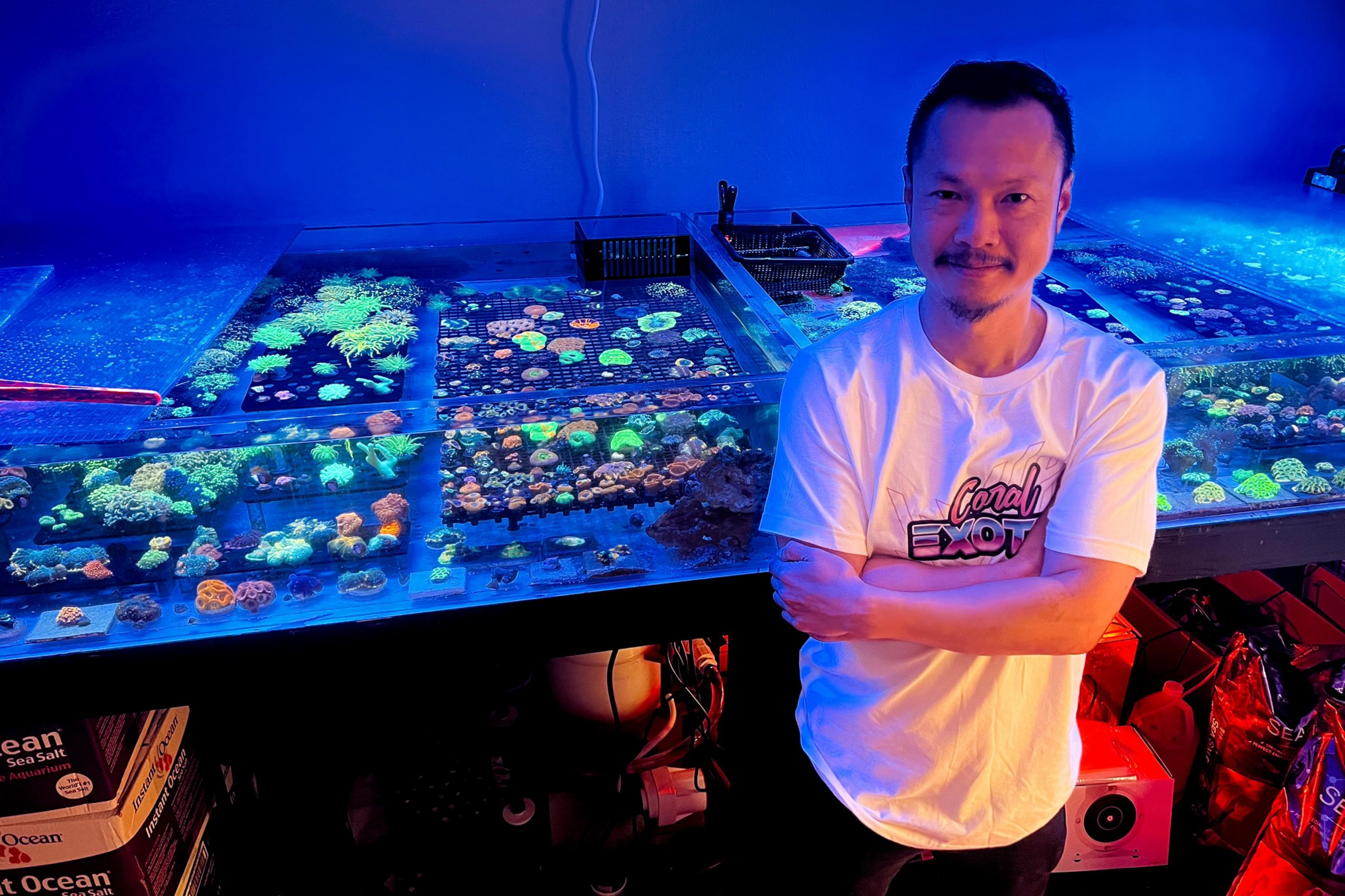 A man smiles, standing beside vibrant coral tanks under blue lighting, arms crossed, wearing a white T-shirt.