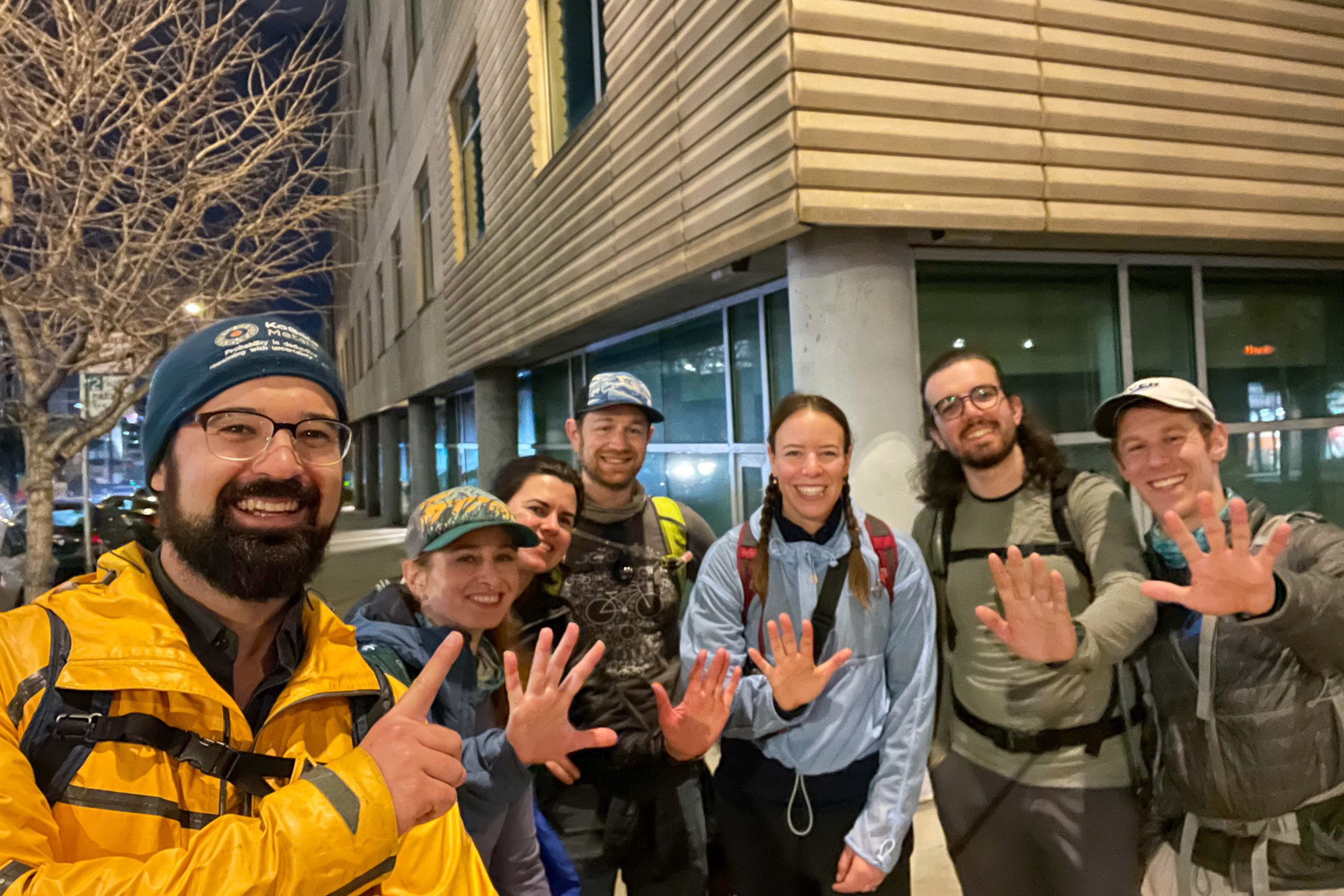 Seven smiling people wearing outdoor gear hold up five fingers, standing close together on a city sidewalk at night near a modern building.
