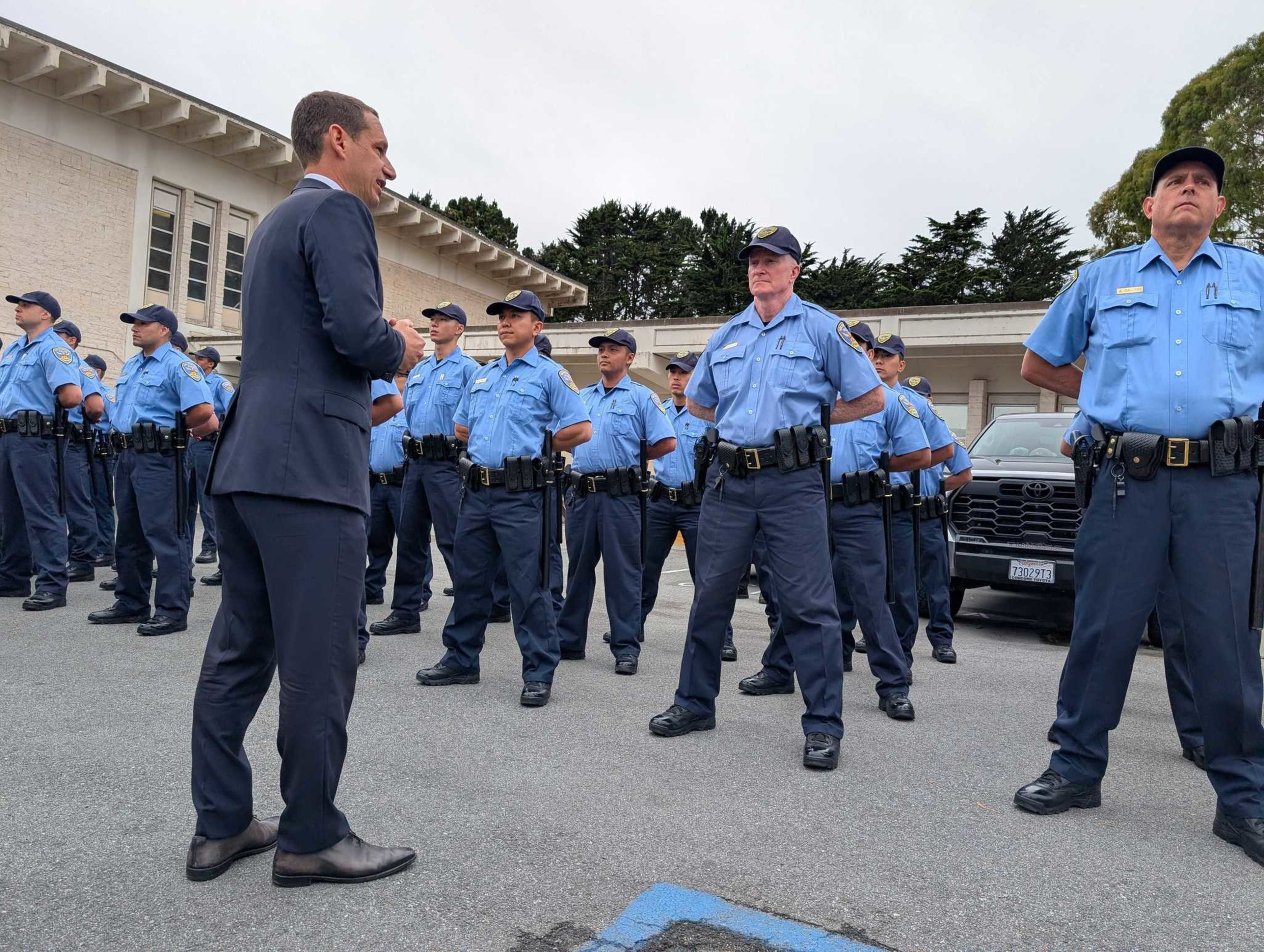 A man in a suit talks to a lined-up group of police officers standing in formation outdoors near buildings and vehicles.