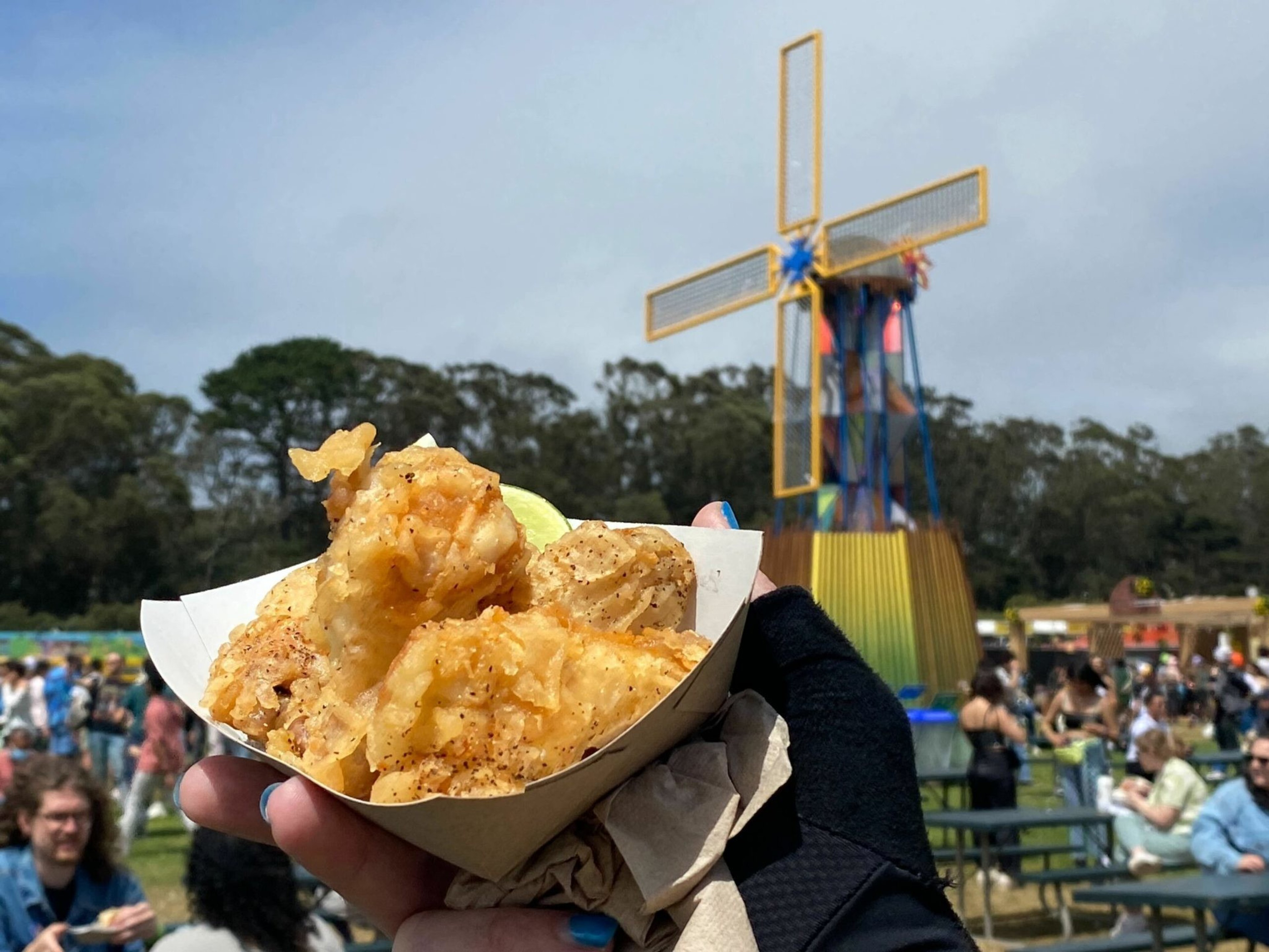 A hand holds a paper tray of crispy fried fish with a lime wedge, against a backdrop of a colorful windmill and a crowded outdoor festival.